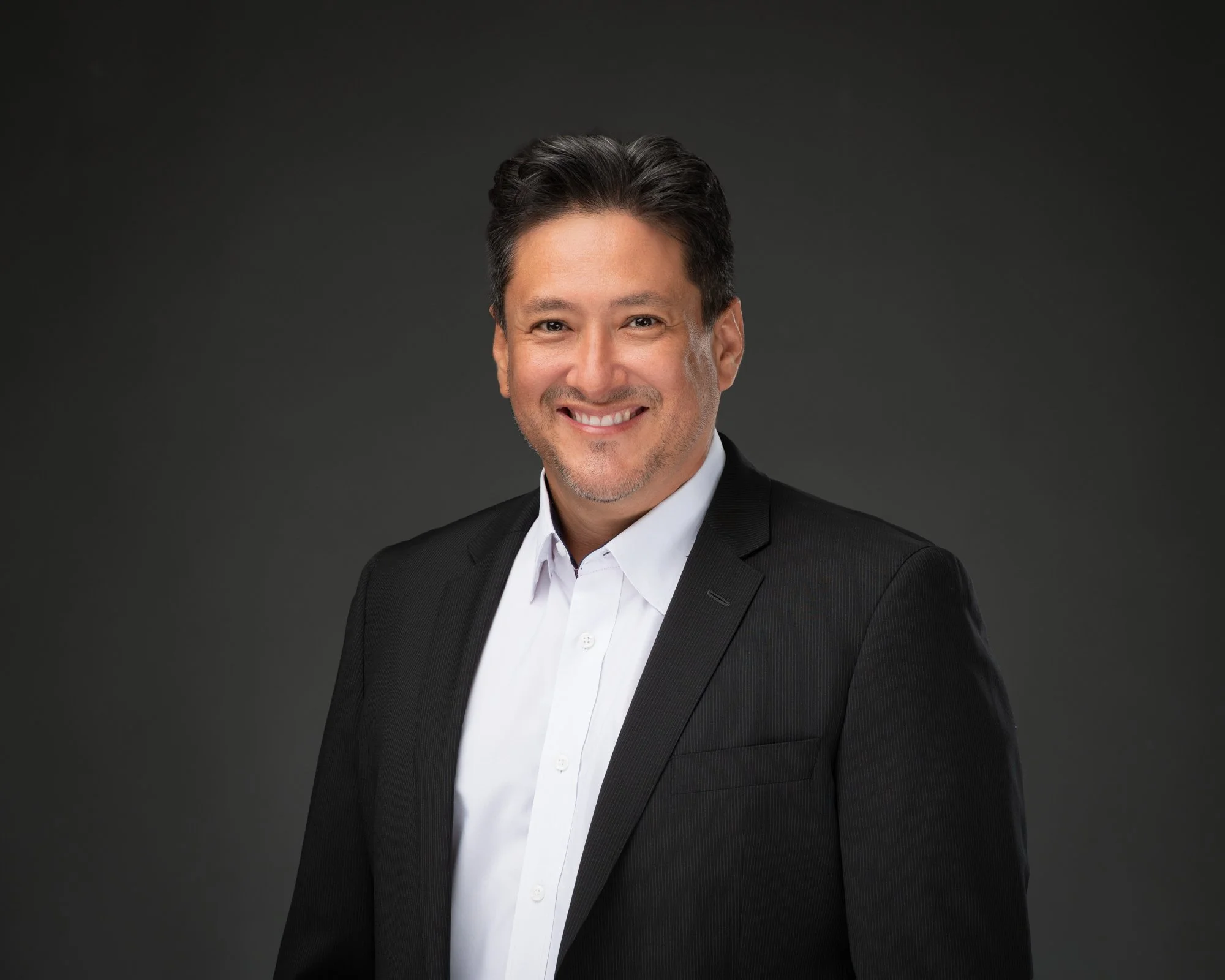 A portrait of a smiling man with dark hair, wearing a black suit and white shirt, against a dark gray background.