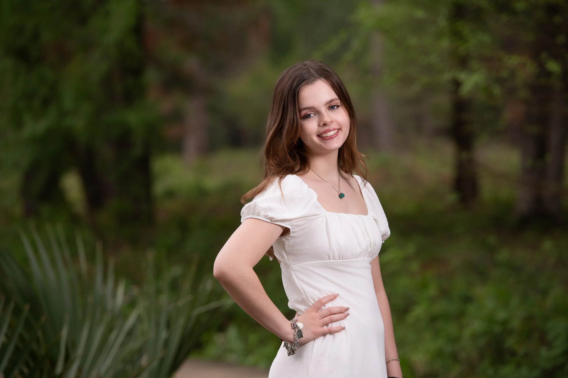 A young woman with shoulder-length brown hair, wearing a white dress and jewelry, stands outdoors with a smile, against a blurred green natural background.