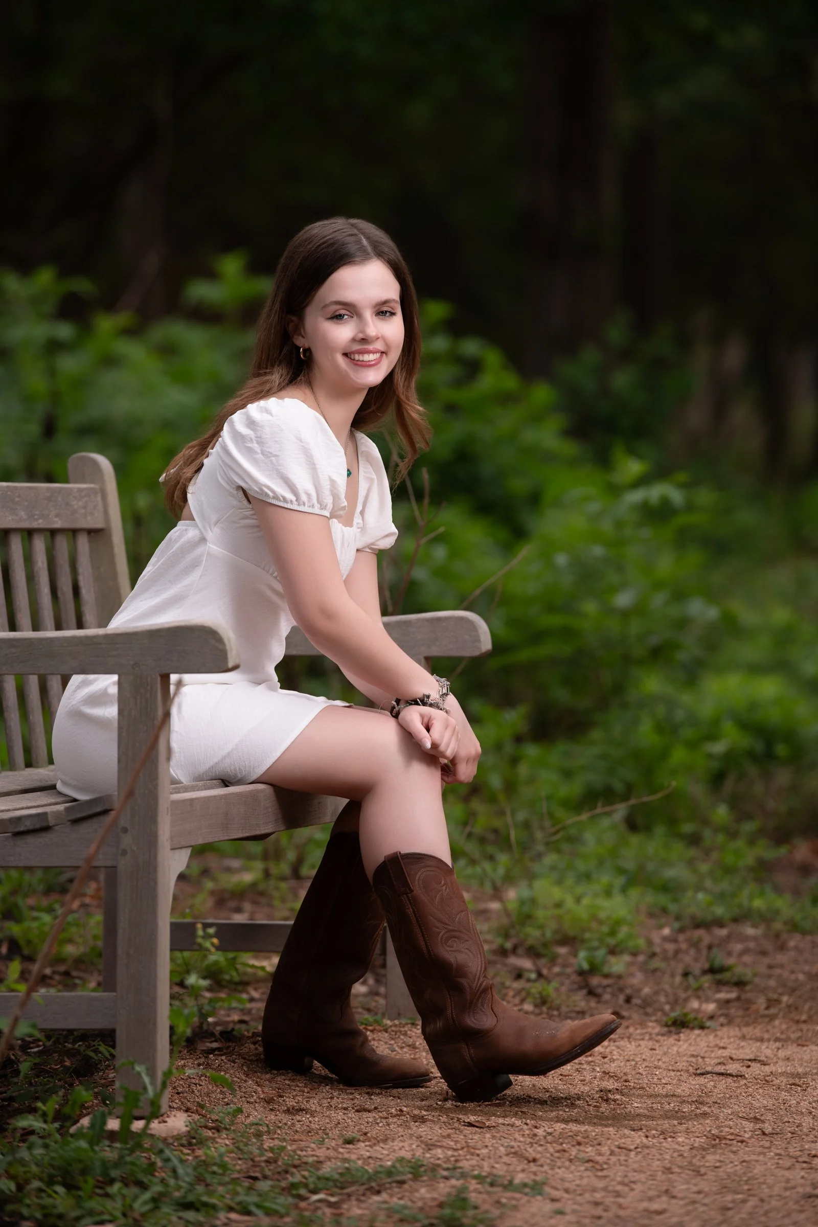 Young woman sitting on a wooden park bench outdoors, smiling at the camera, wearing a white dress and brown cowboy boots, with green foliage and trees in the background.