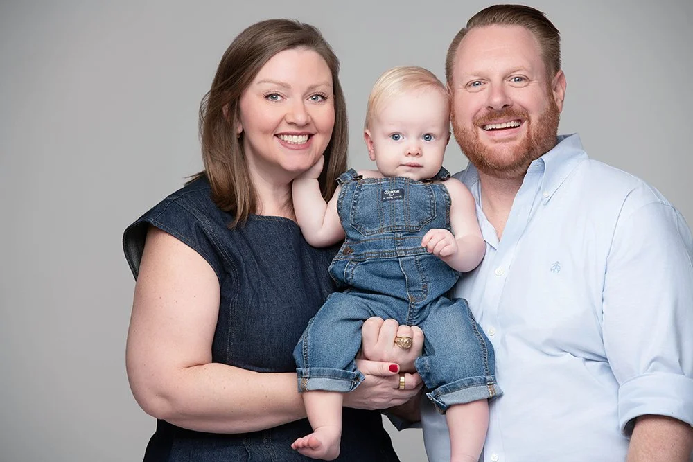 A smiling woman, smiling man, and a small child with blonde hair in denim overalls against a plain gray background.