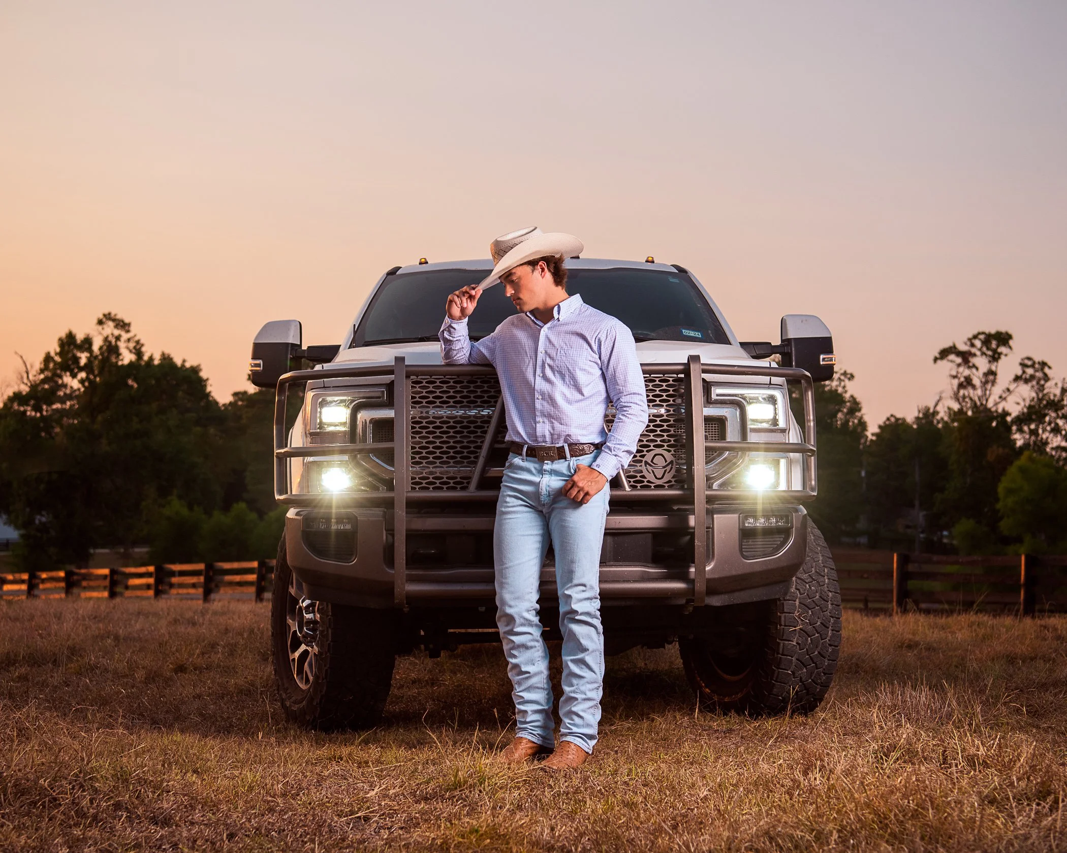 A man in a cowboy hat and button-up shirt standing in front of a large pickup truck on a grassy field at sunset.