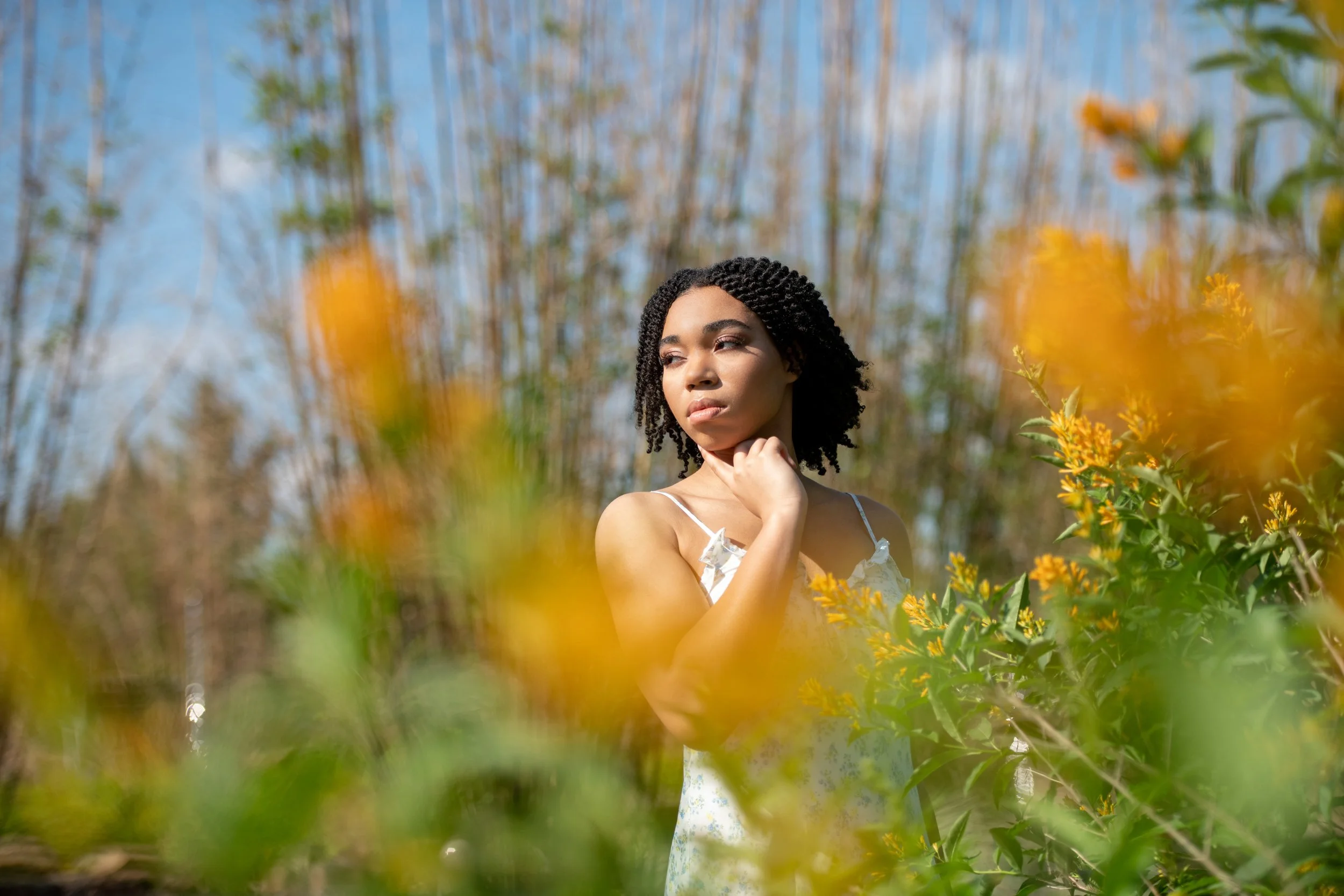 A woman with dark curly hair and light skin standing among yellow flowers outdoors, with blue sky and trees in the background.