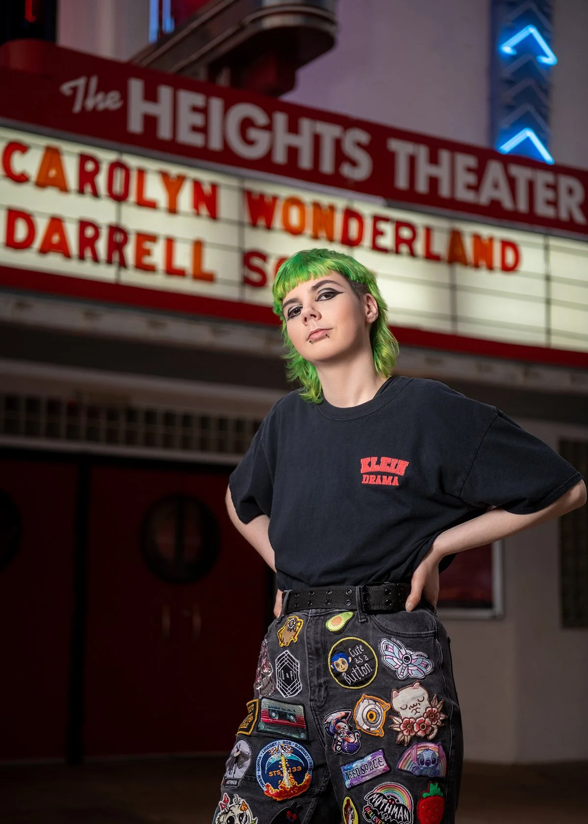 Person with short green hair and gothic makeup standing with hands on hips in front of a theater marquee advertising 'The Heights Theater,' with a woman in a black oversized t-shirt covered in patches.