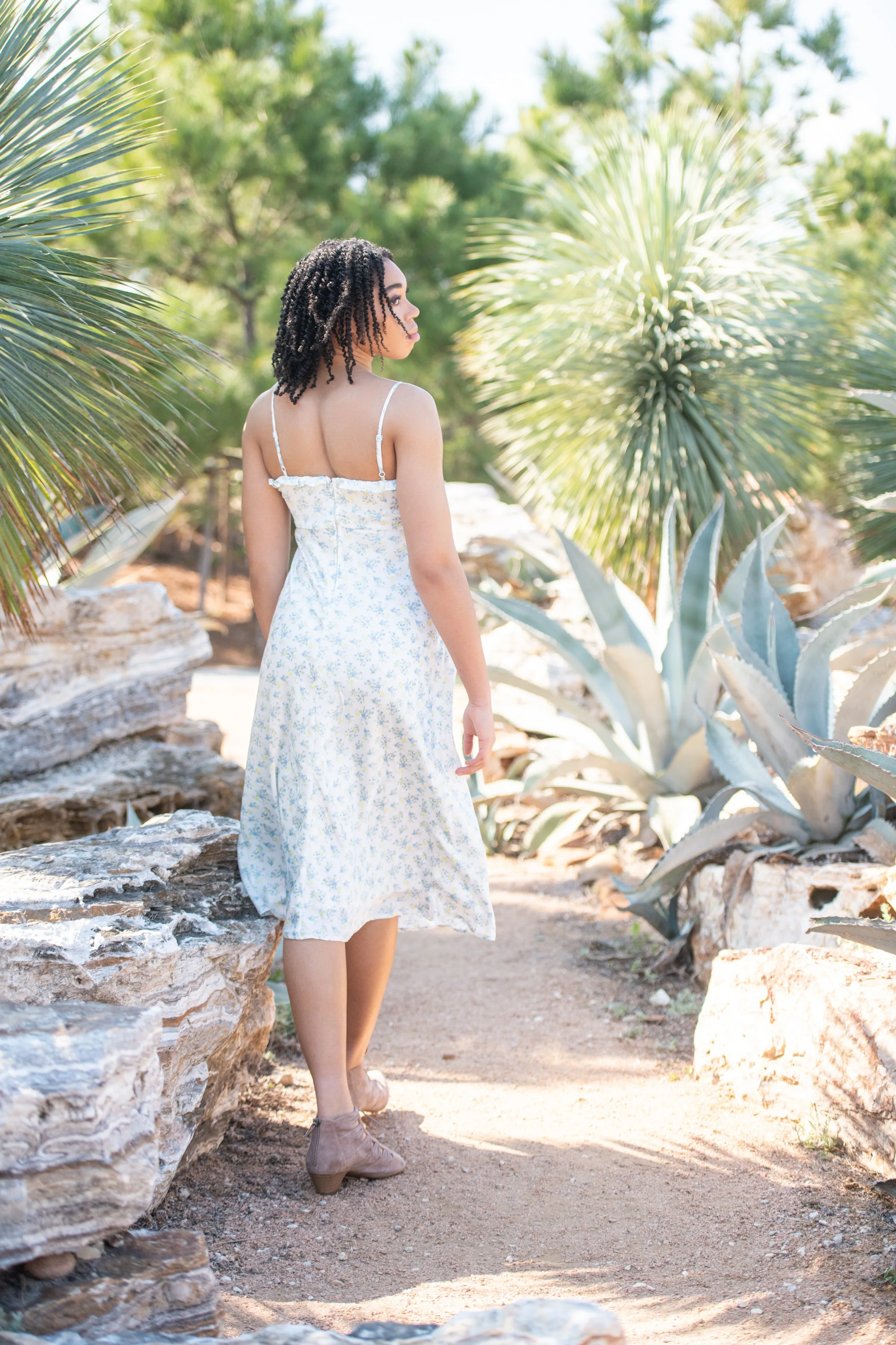 A woman with dark, curly hair wearing a white and yellow floral dress, standing barefoot among desert plants and rocks, looking to her right in a sunny outdoor setting.