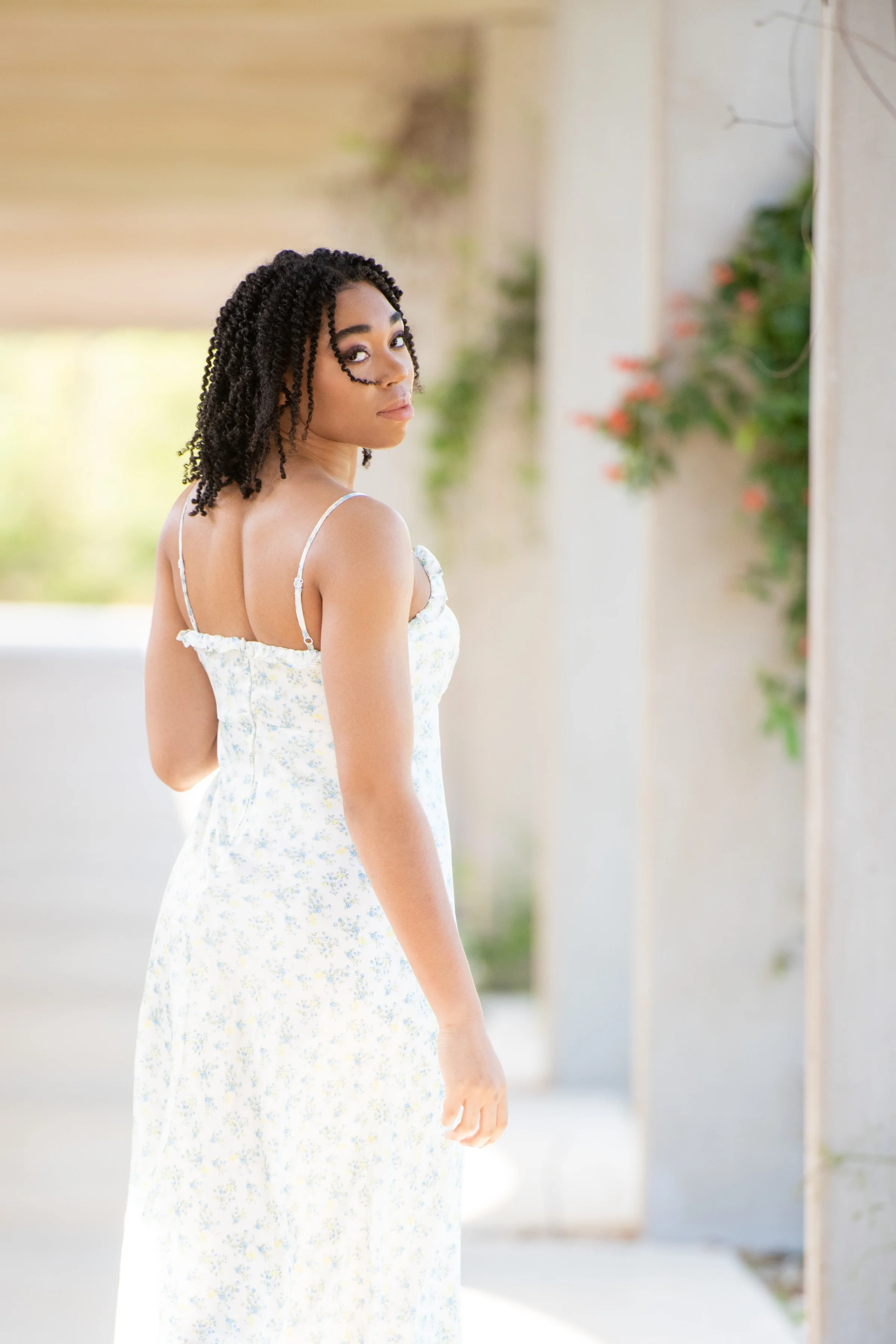 A young woman with dark curly hair in a white sundress with a small floral pattern, standing outdoors and looking over her shoulder at the camera. She is in front of a beige wall with a garden in the background.