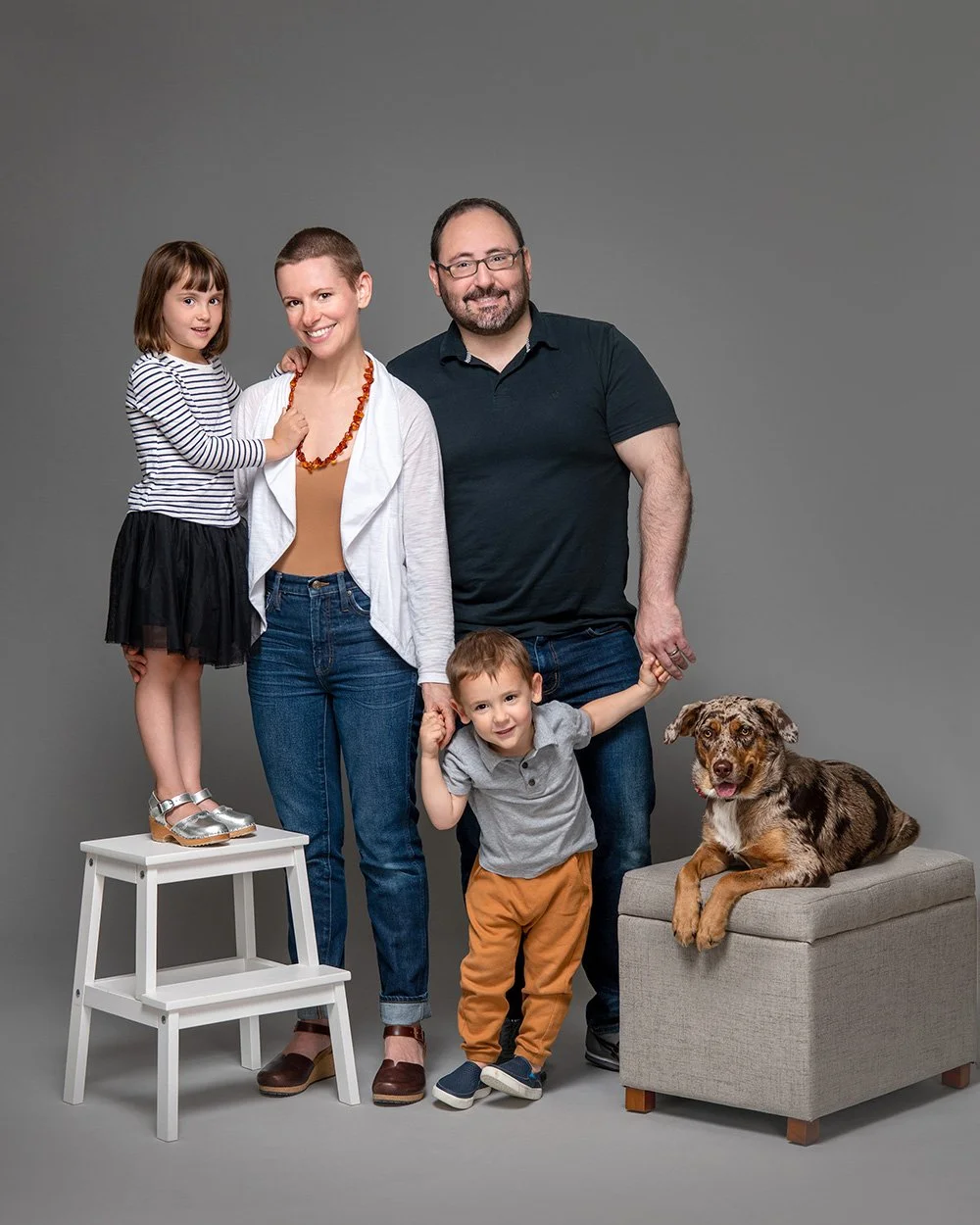 Family portrait of two adults, two children, and a dog against a gray background. The woman and girl stand on the left, the man and boy on the right. The girl is on a white step stool, and the dog is sitting on a small gray ottoman.