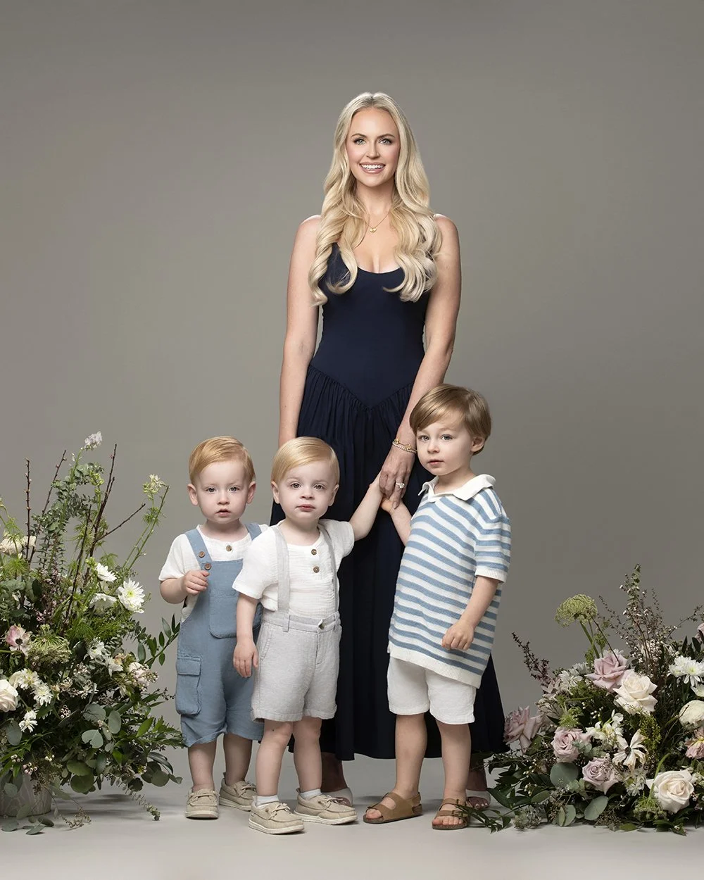 A woman with long blonde hair wearing a dark blue dress standing behind three young boys, all surrounded by floral arrangements, posing against a gray background.