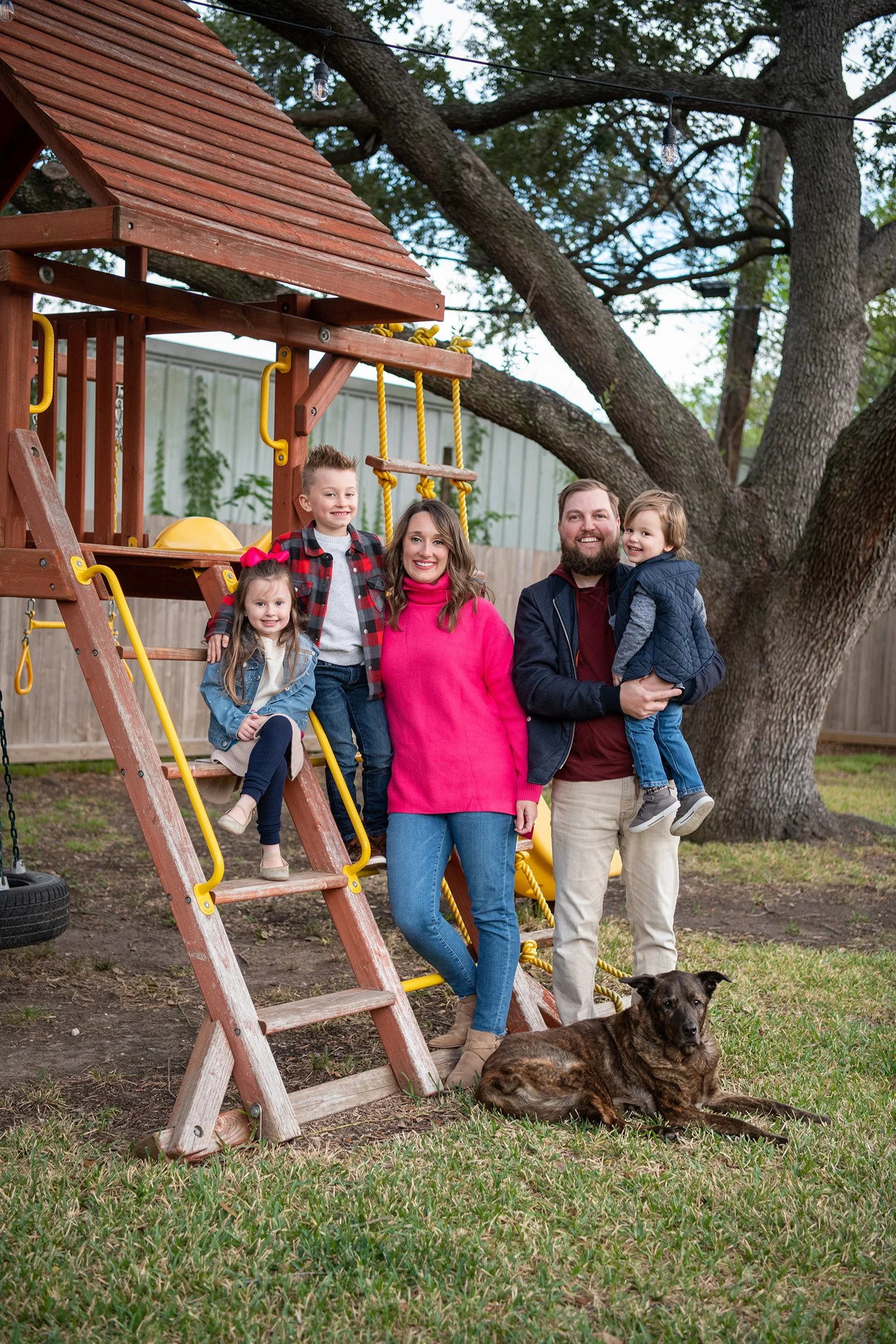 A family with children and a dog standing near a wooden play structure in a backyard with large trees and string lights.
