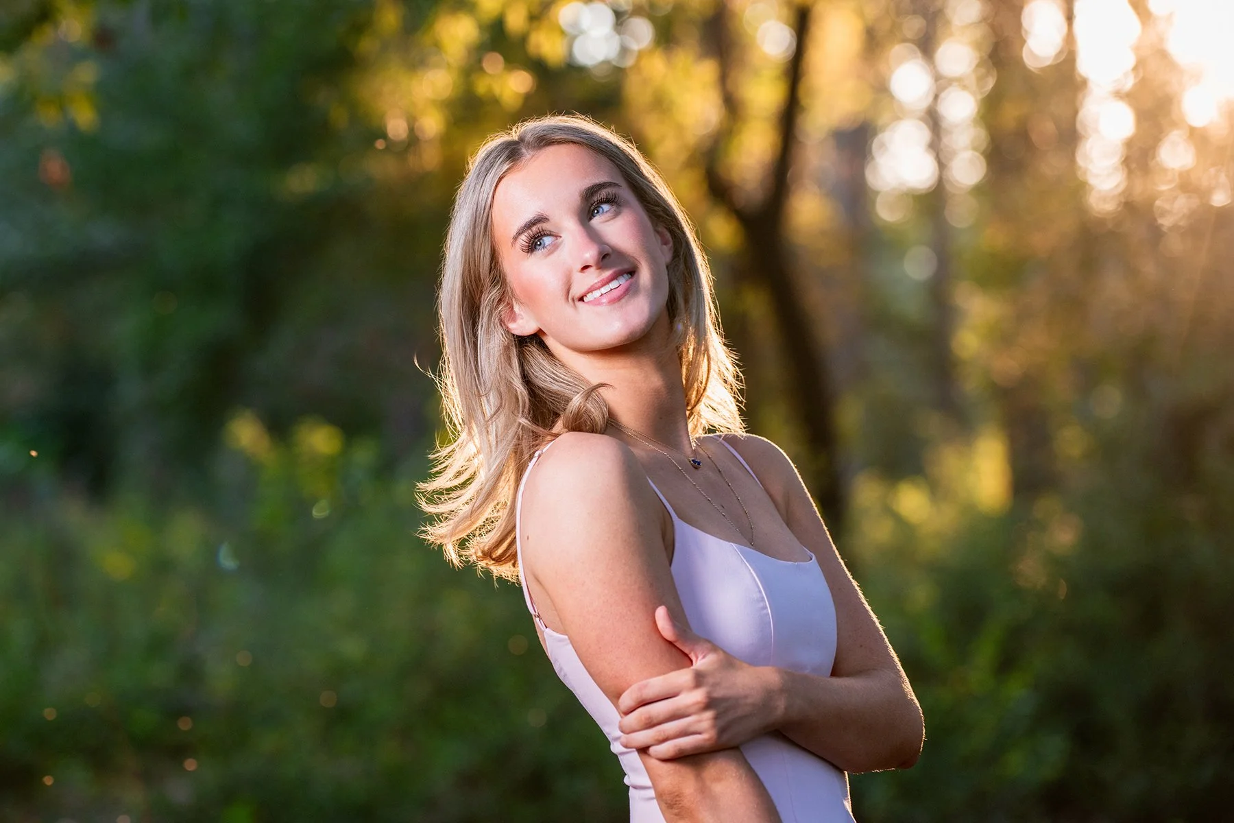 A young woman with blonde hair and blue eyes smiling outdoors in a park with sunlight filtering through the trees, wearing a white sleeveless top and a necklace.
