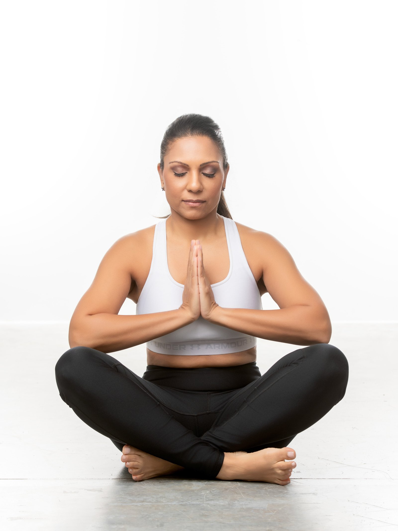 A woman practicing yoga or meditation in a seated cross-legged position, with her hands pressed together in a prayer gesture and eyes closed, on a white background.