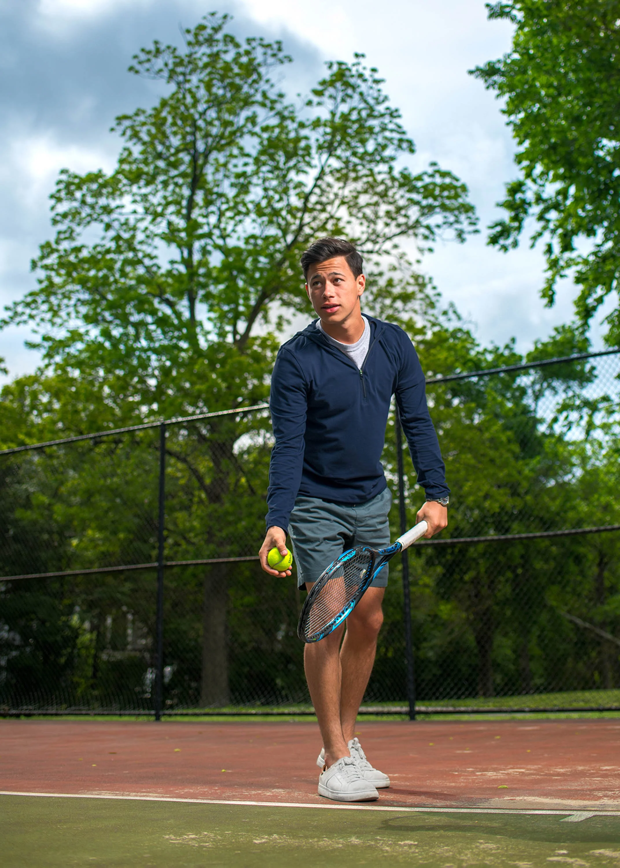 A young man in a navy blue jacket, gray shorts, and white sneakers on a tennis court, holding a tennis ball and racket, with trees and a cloudy sky in the background.