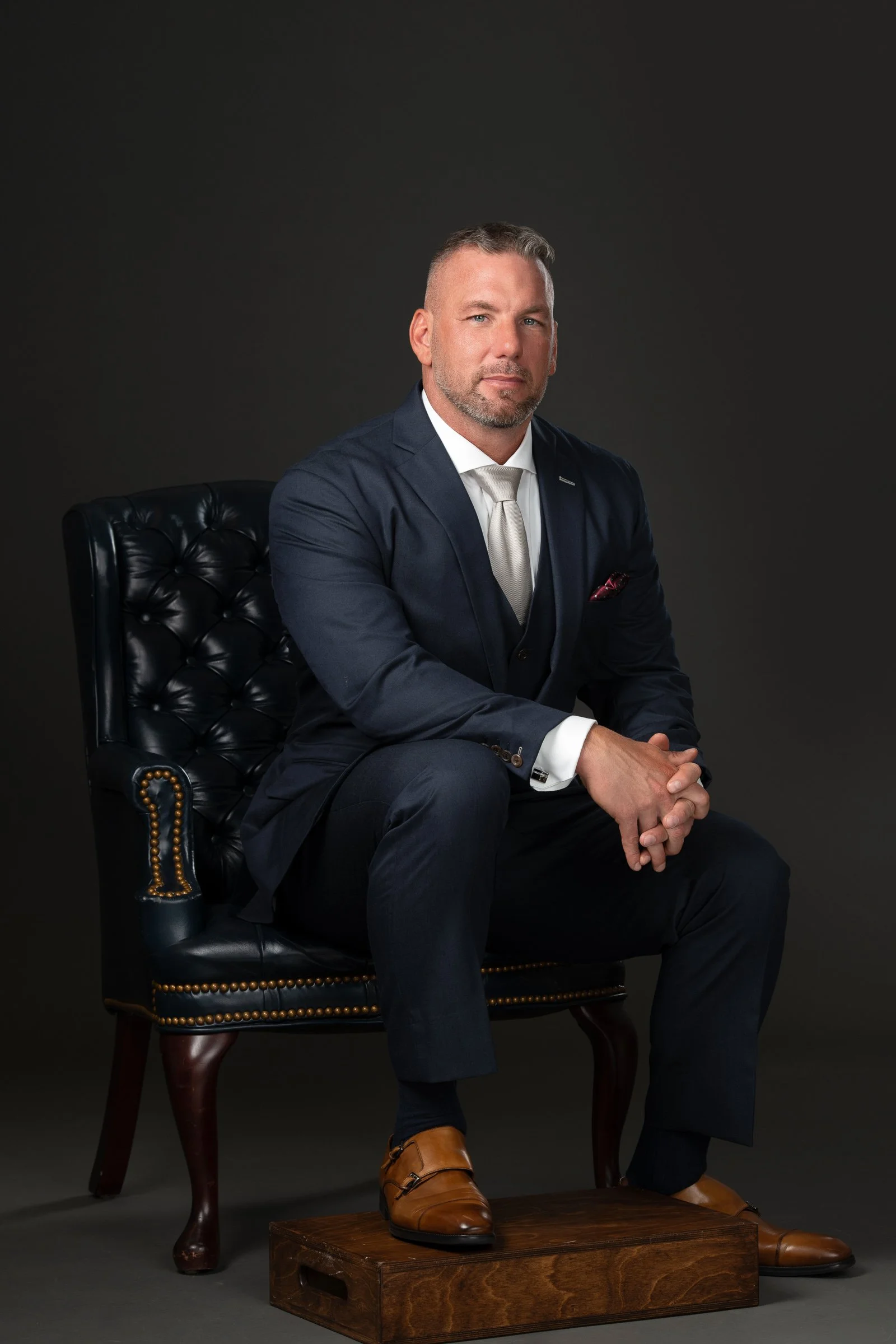 A man in a navy suit and light tie seated on a black tufted leather armchair, with a dark background, wearing brown shoes and resting his hands on his knee a wooden block.