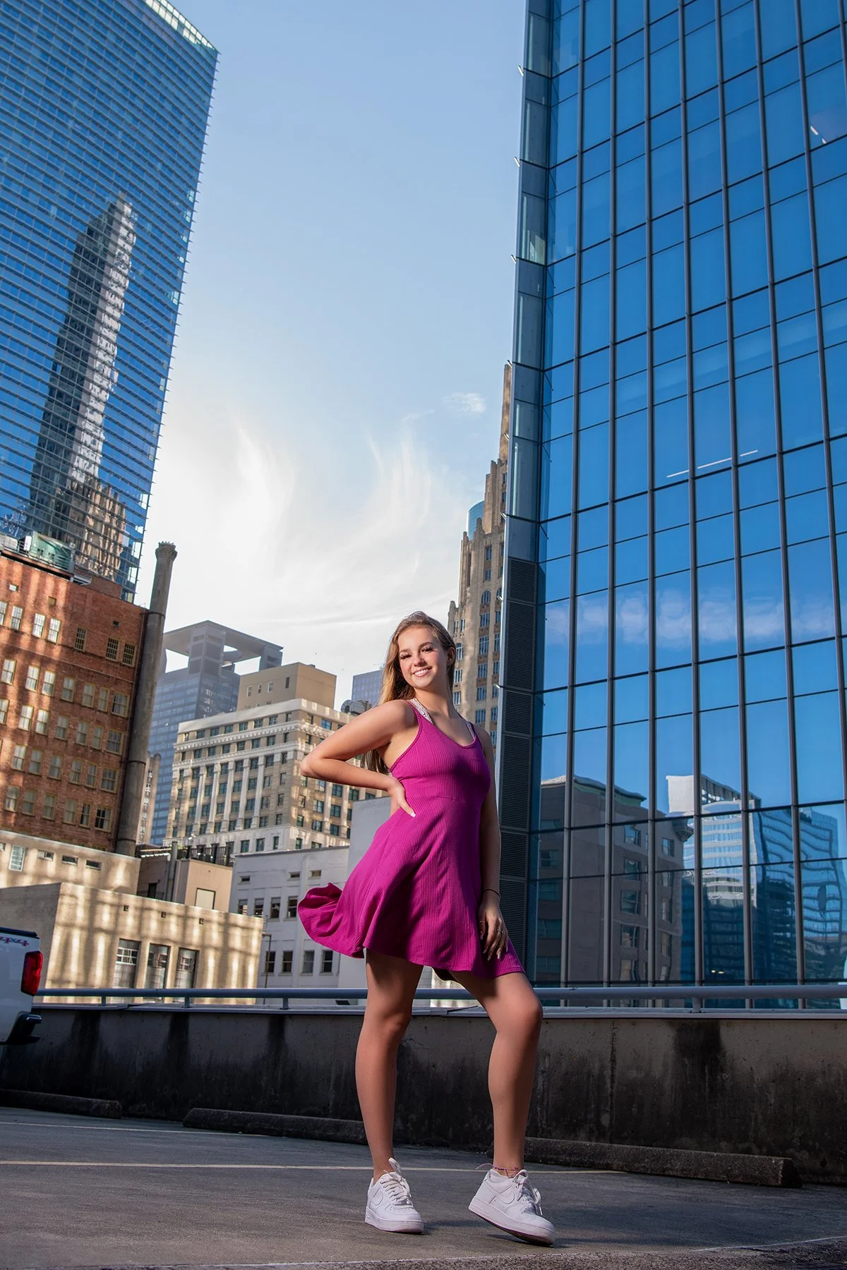 A young woman in a bright pink dress and white sneakers posing in an urban cityscape with tall glass buildings and a blue sky.