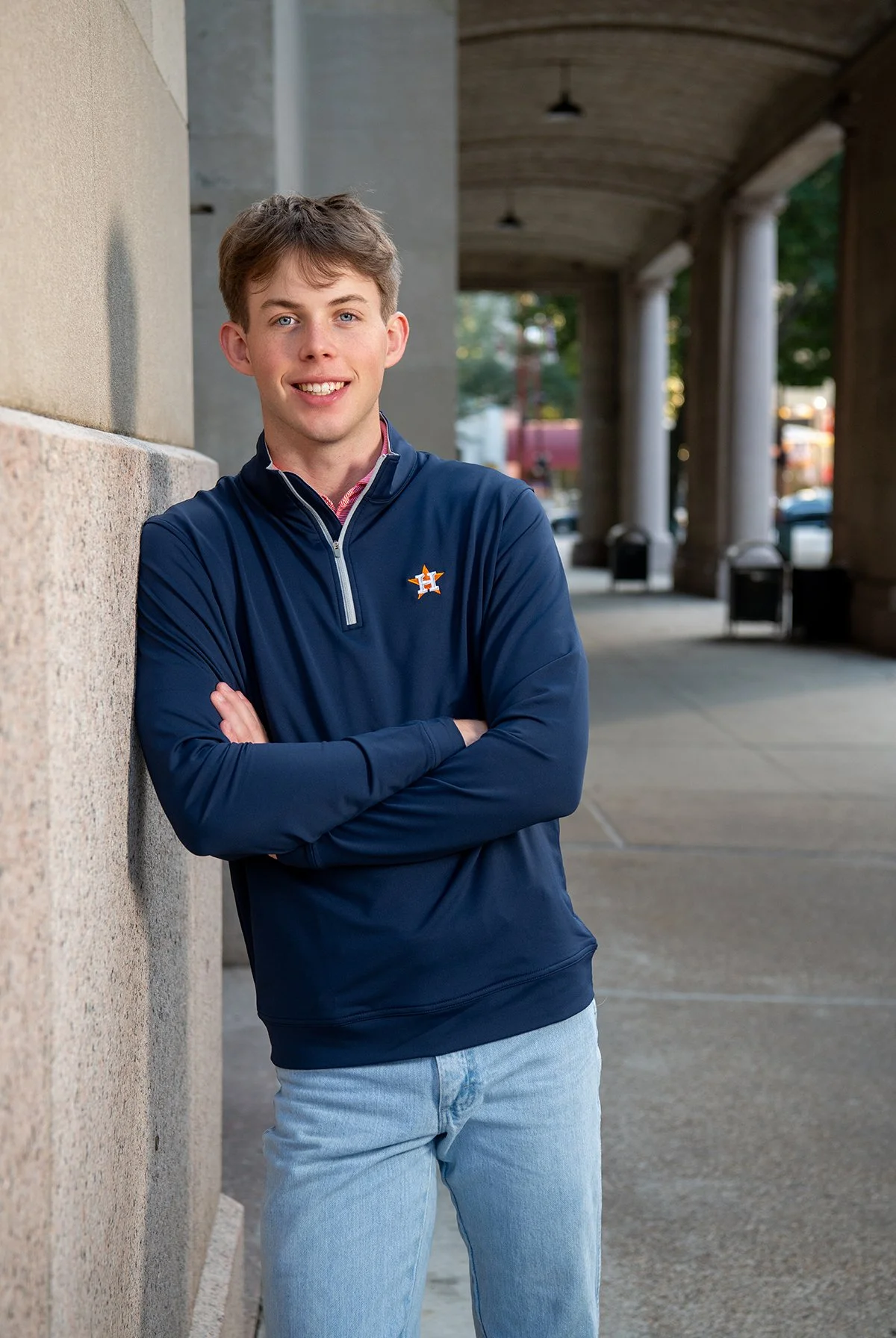 Young man with light brown hair wearing a navy Houston Astros jacket and light blue jeans standing with arms crossed outdoors.