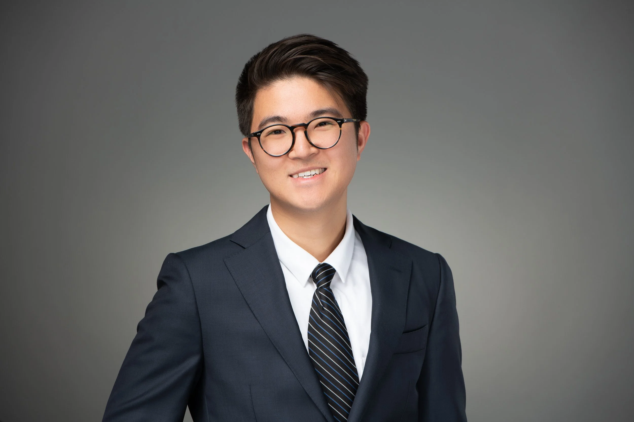 Professional young man in suit and tie smiling, wearing glasses, against a plain gray background.