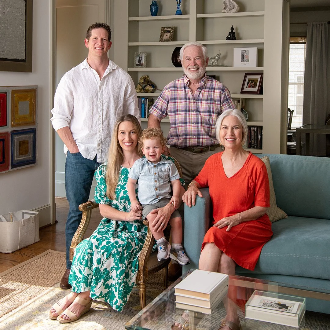 A multigenerational family of six posing together in a living room. Four adults and two children, one sitting on a chair and the other on the sofa. They are smiling at the camera, with a bookshelf and artwork in the background.