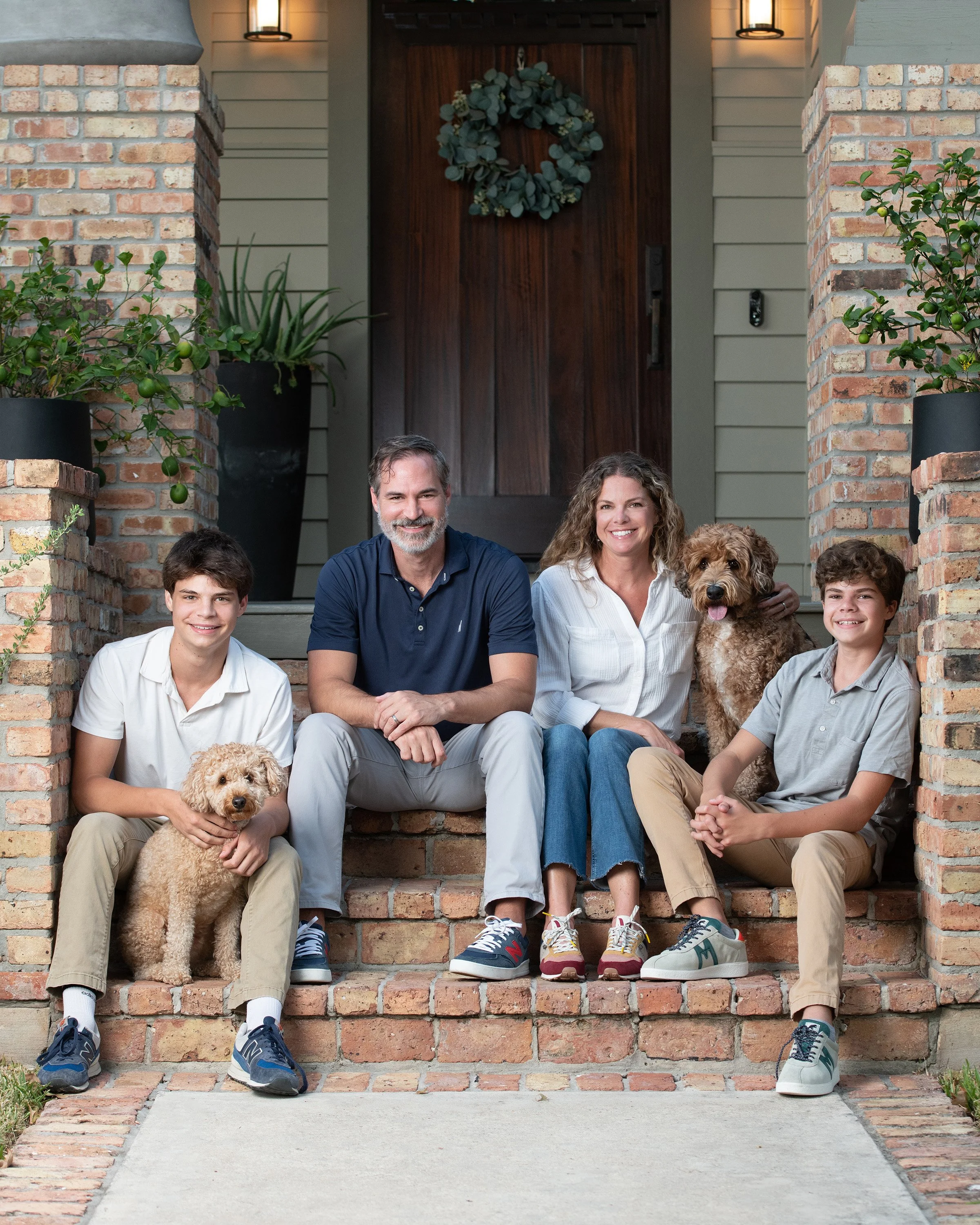 A family sitting on front steps of a house, smiling, with a dog. The house has brick columns, green plants, and a wreath on the door.