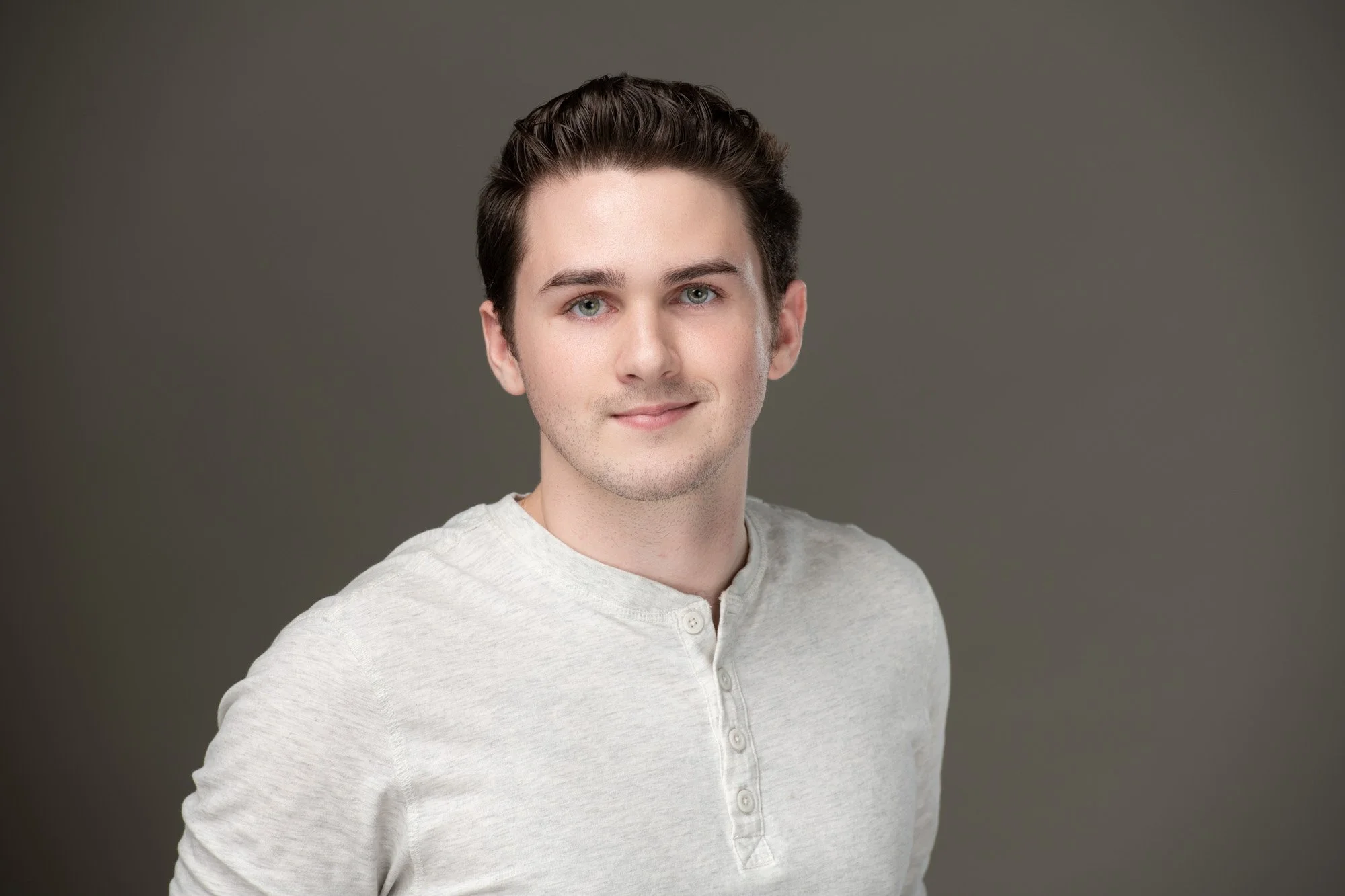 Headshot of a young man with dark hair, light green eyes, wearing a light gray henley shirt, smiling softly, against a plain gray background.
