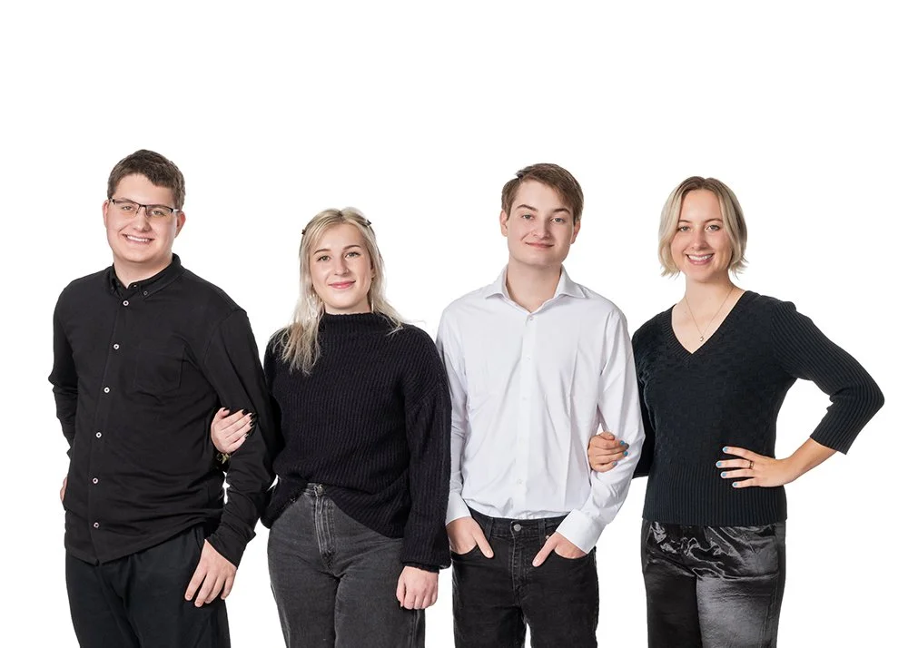 Four young adults standing together against a plain white background, smiling and looking at the camera.