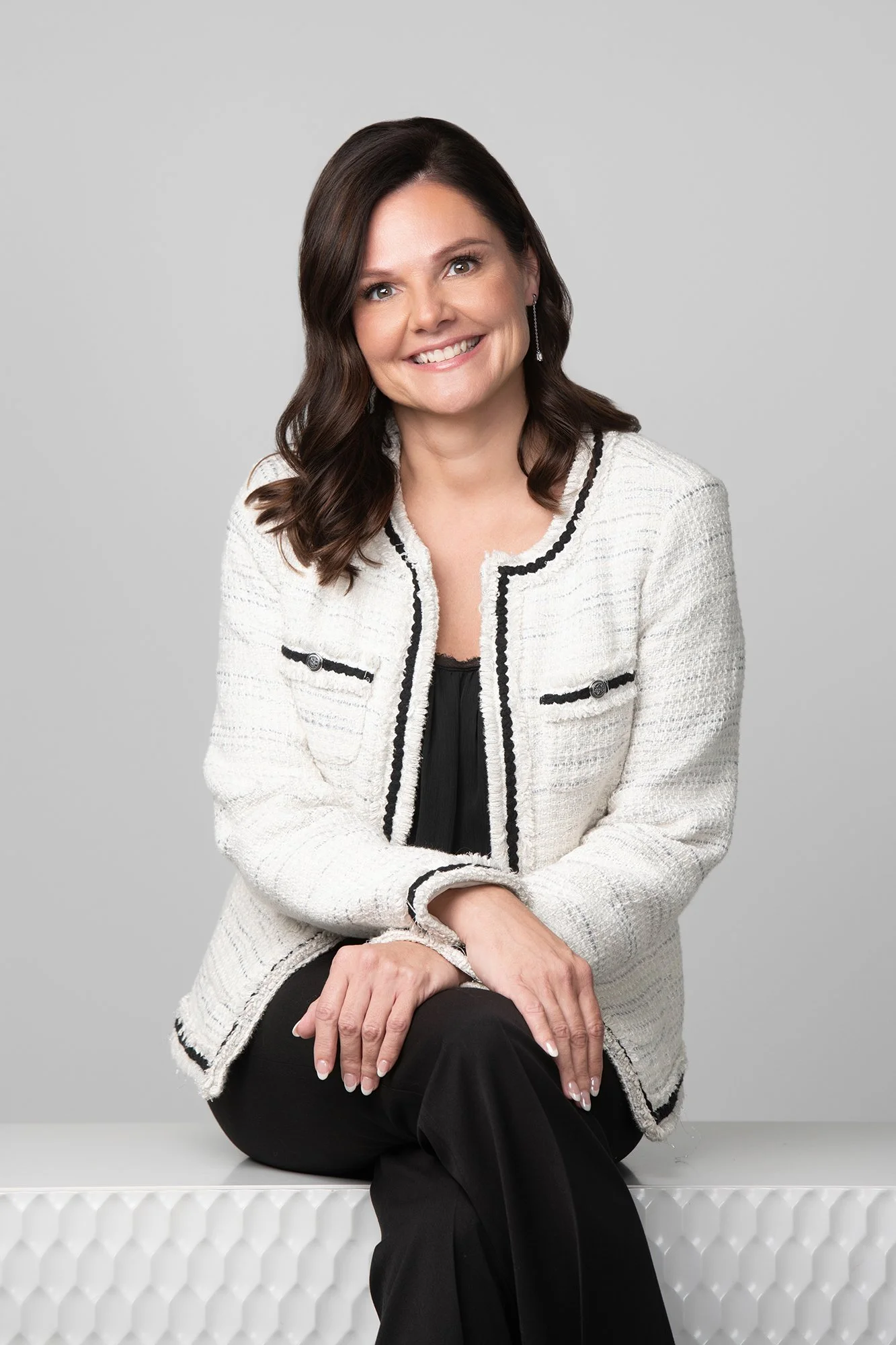 A woman with shoulder-length dark brown hair styled in loose waves, smiling and sitting on a white textured surface against a plain light gray background. She is wearing a white textured blazer with black trim, a black top, and black pants. She has d