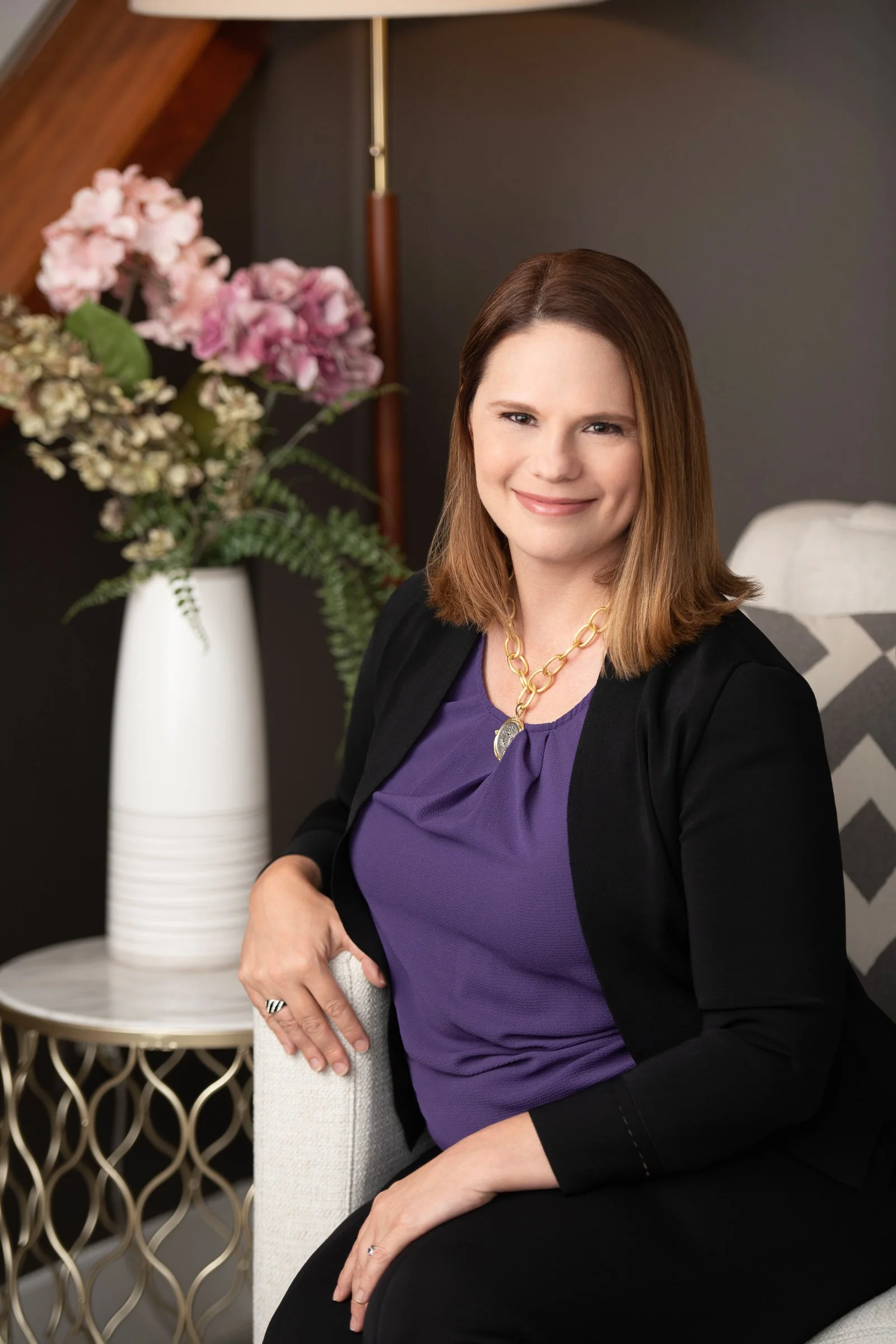A woman with shoulder-length brown hair smiling, sitting on a beige armchair, wearing a purple blouse, black blazer, black pants, gold necklace, and a ring on her left hand. Behind her is a white vase with pink and white flowers on a small round tabl