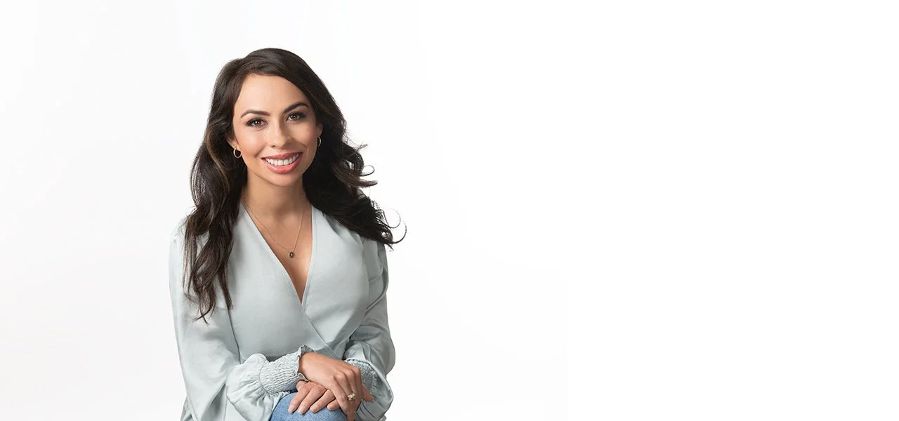 A woman with long, dark hair and a bright smile, wearing a light gray blouse and jewelry, sitting against a white background.