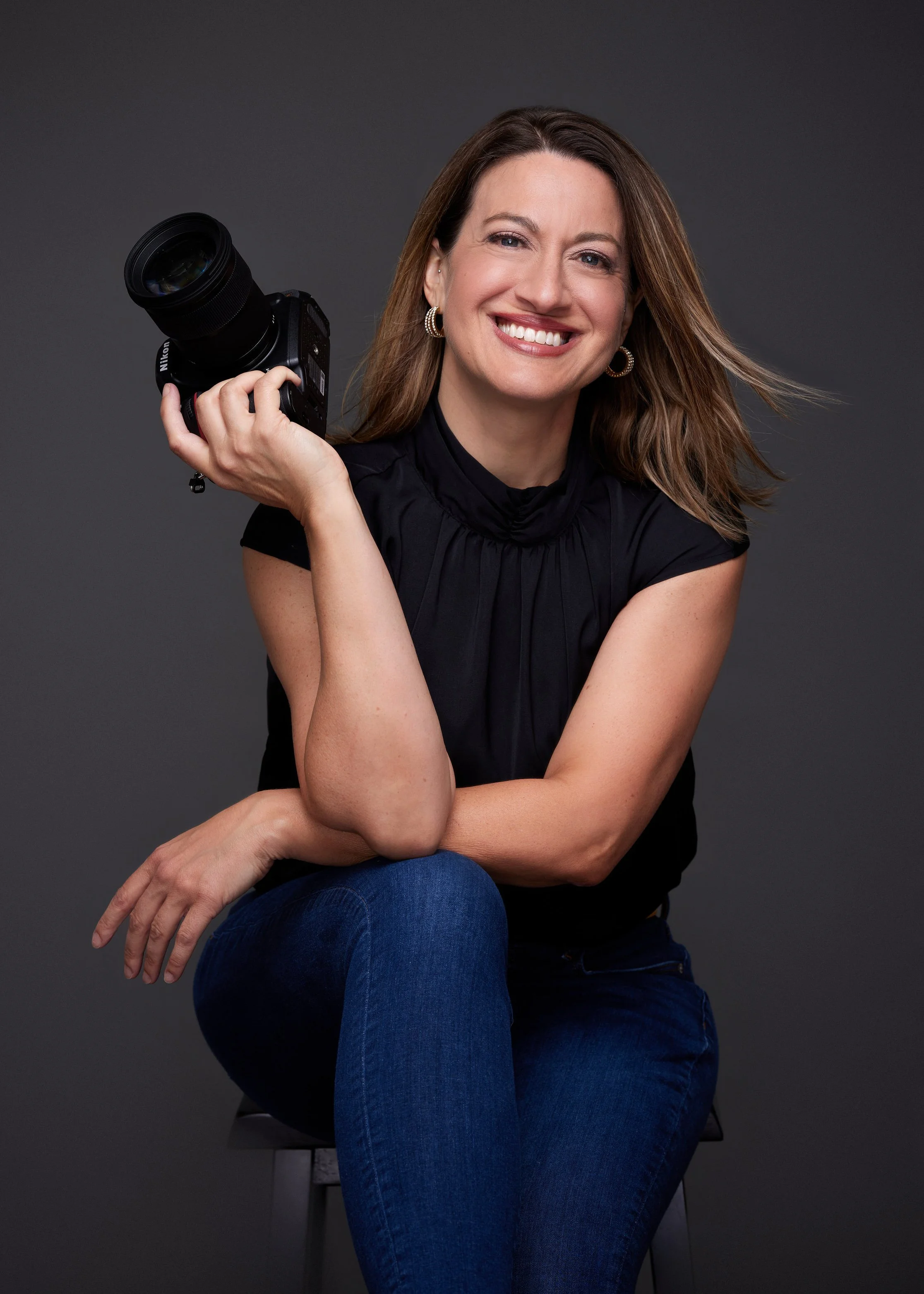 Smiling woman with shoulder-length brown hair, wearing earrings and a black top, sitting on a chair against a dark background, holding a professional camera in her right hand.