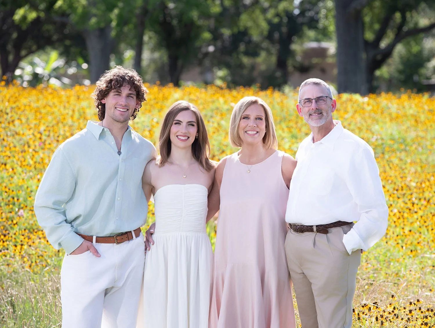 A family of four standing outdoors in front of a field of yellow flowers, smiling at the camera, on a sunny day.