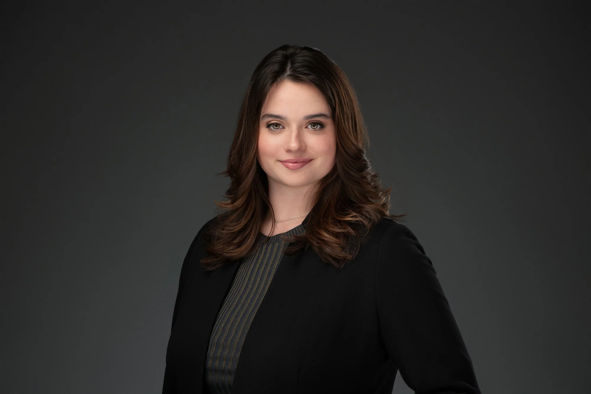 Portrait of a young woman with long, wavy brown hair, wearing a black blazer over a dark striped shirt, standing against a dark gray background, smiling subtly.
