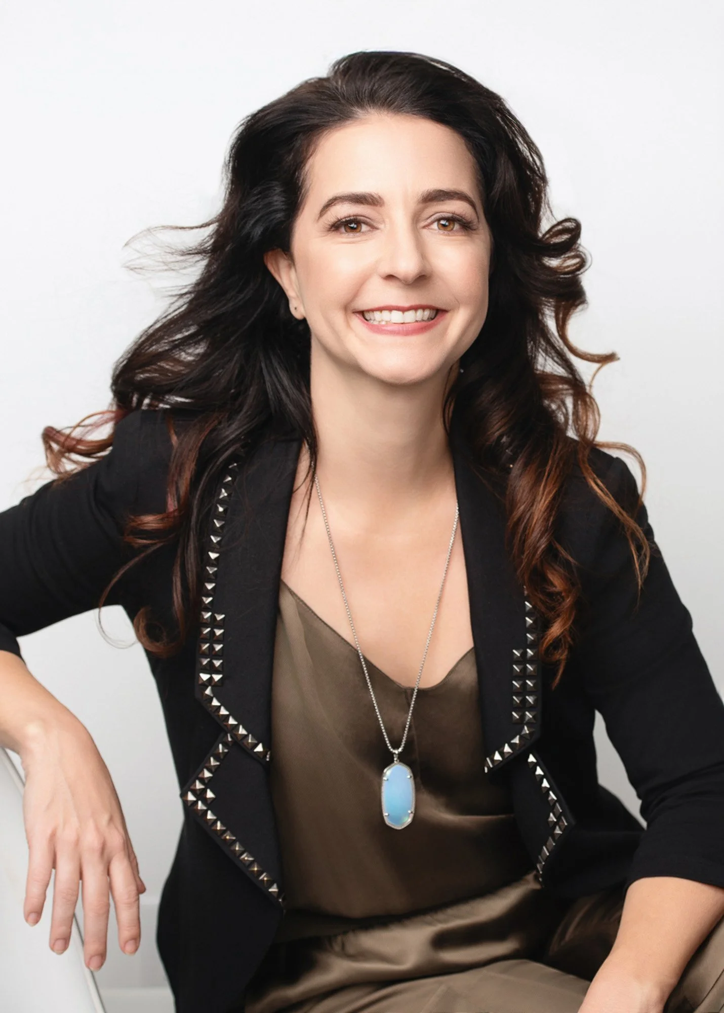 Woman with brown hair and brown eyes smiling, wearing a black blazer with studded trim, a taupe top, and a long necklace with a large oval pendant, seated against a plain white background.
