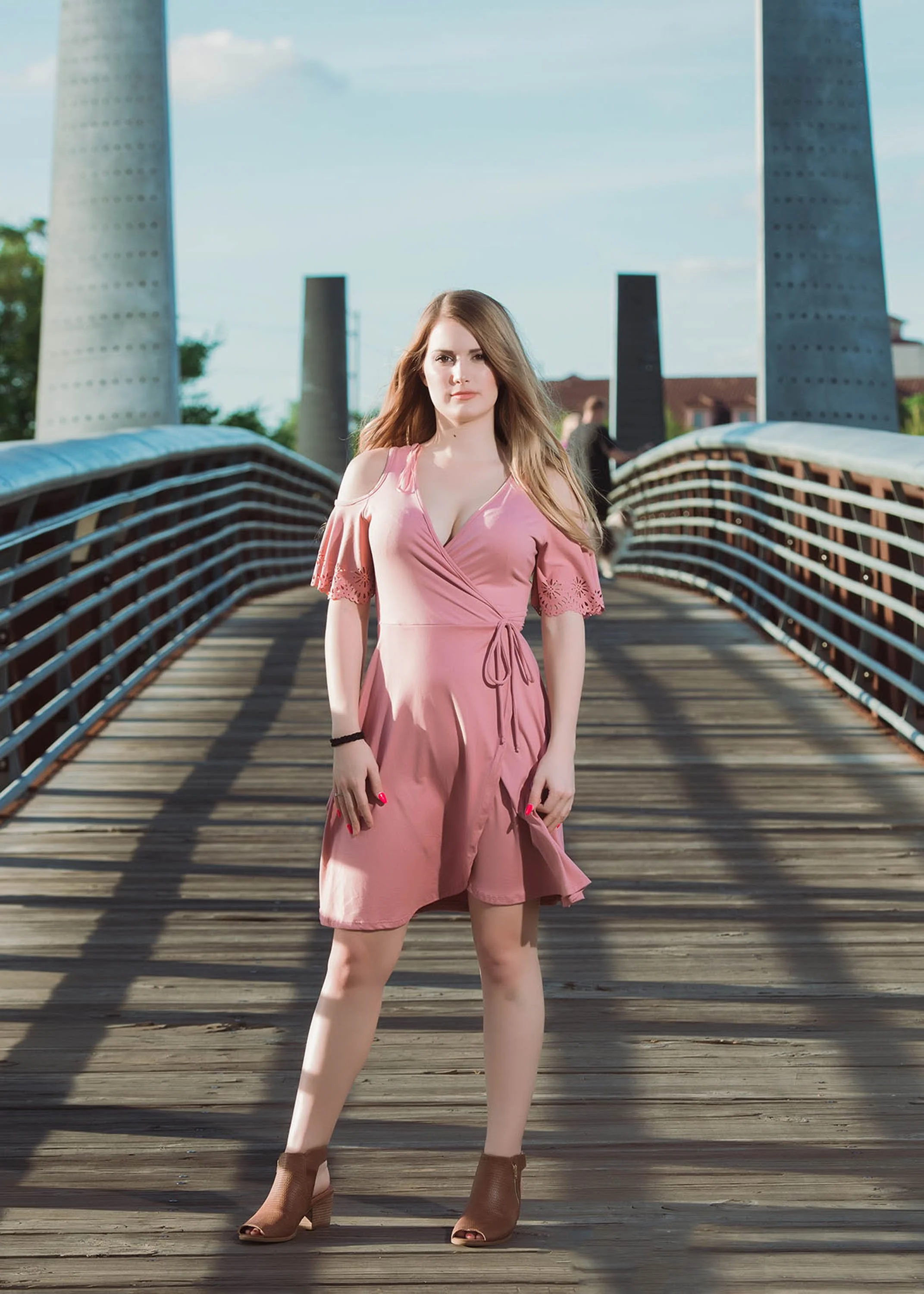 Young woman in a pink dress standing on a wooden bridge with large towers in the background.