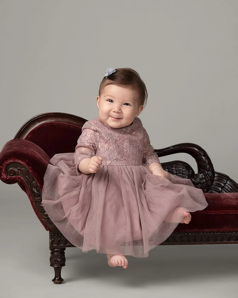 A smiling baby girl in a pink dress sitting on an antique dark wood and red velvet chaise lounge.