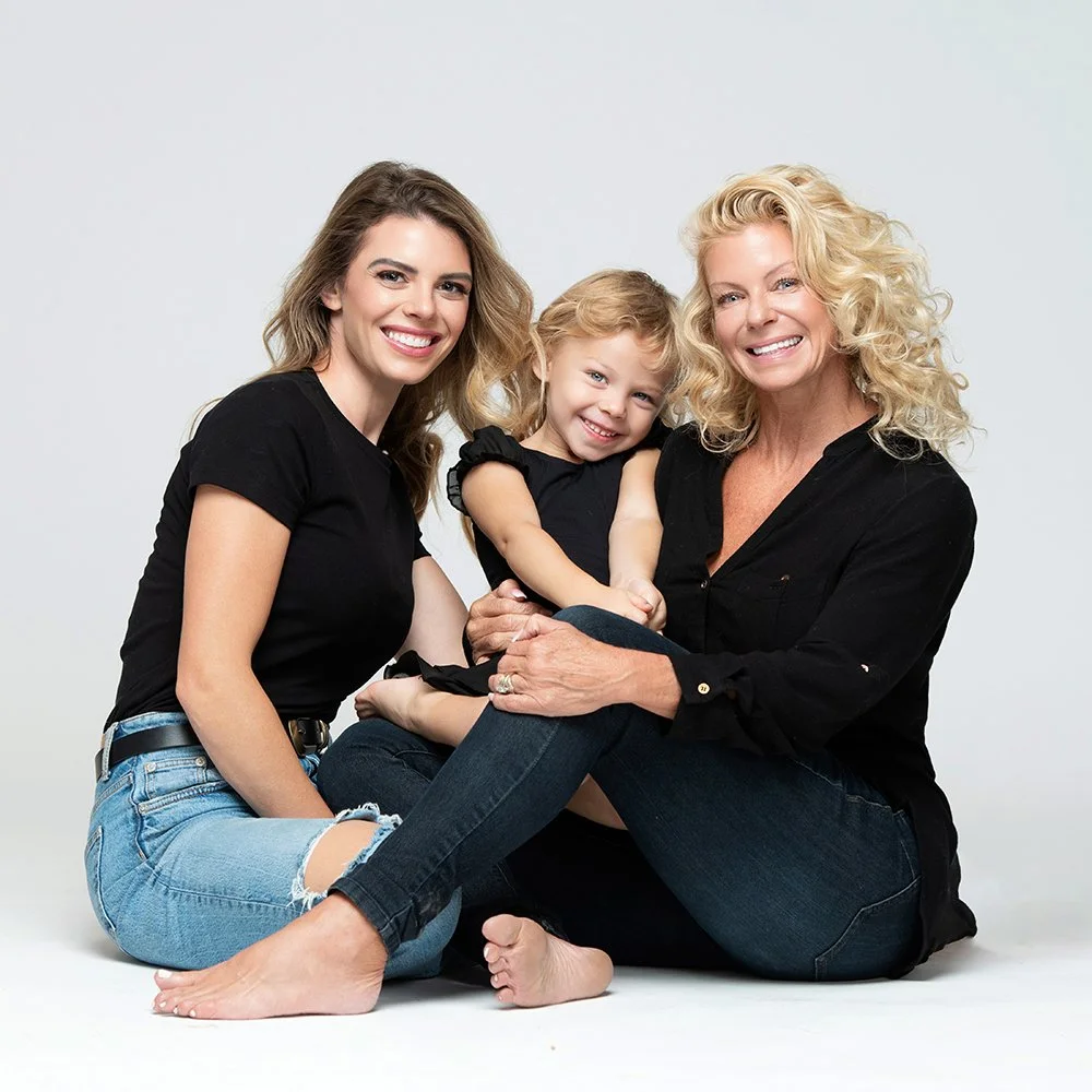 Three females, a young woman, a little girl, and an older woman, sitting together on the floor, smiling, with the little girl sitting on the older woman's lap and the young woman next to them.