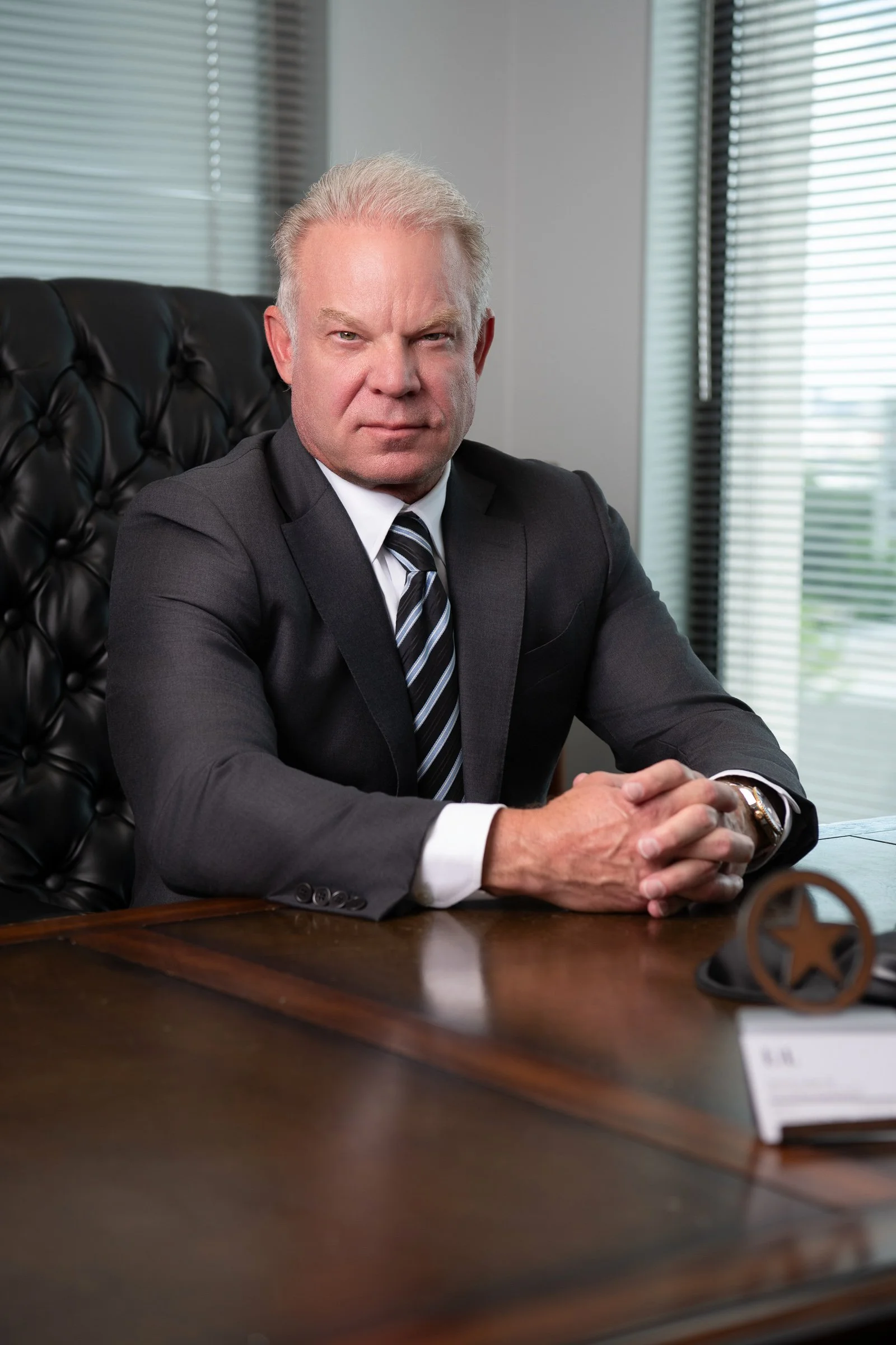 A middle-aged man with gray hair and a serious expression, sitting at a large wooden desk in an office with large windows and horizontal blinds, wearing a dark suit and striped tie.