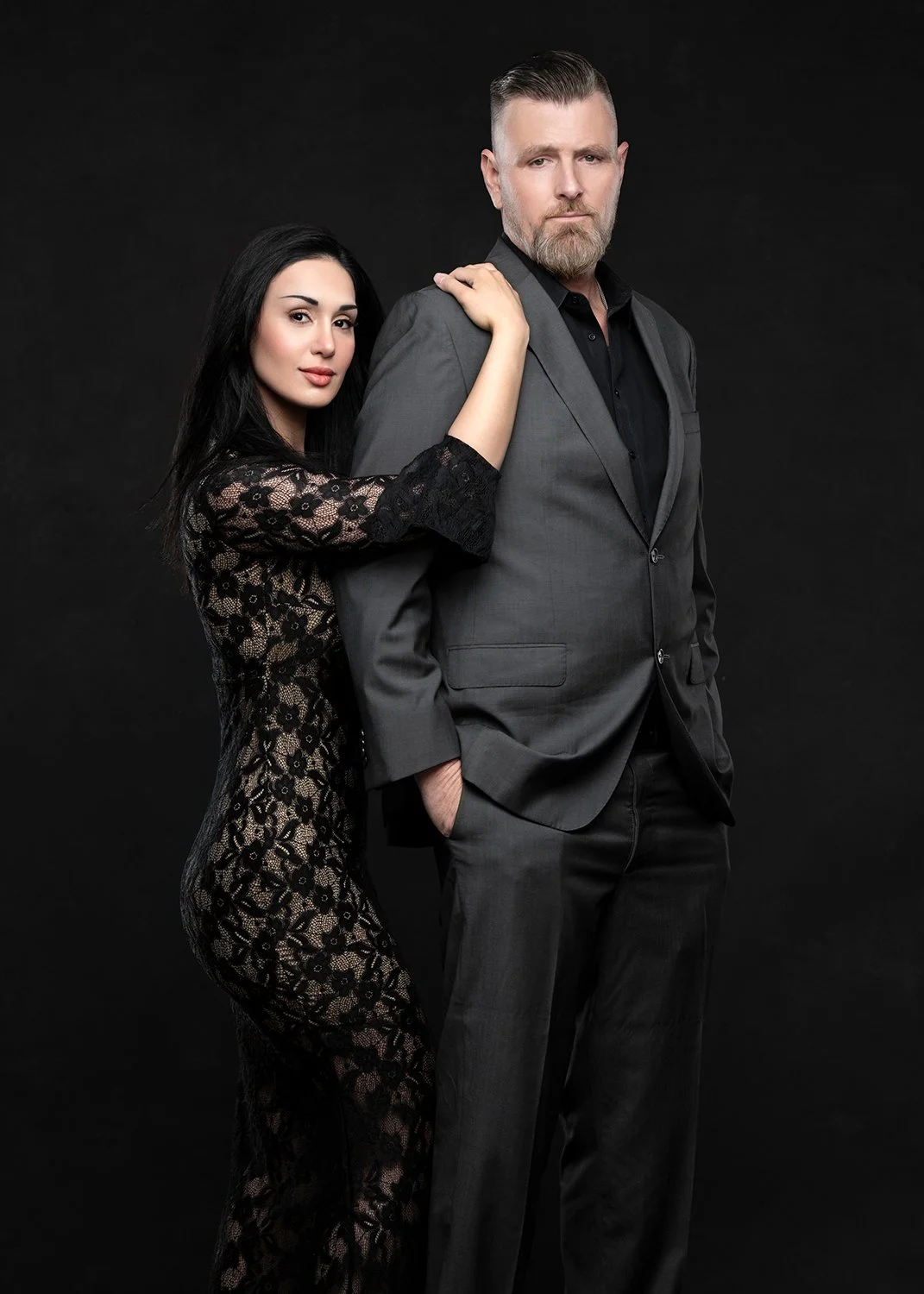 A woman in a black lace dress standing next to a man in a gray suit against a dark background.