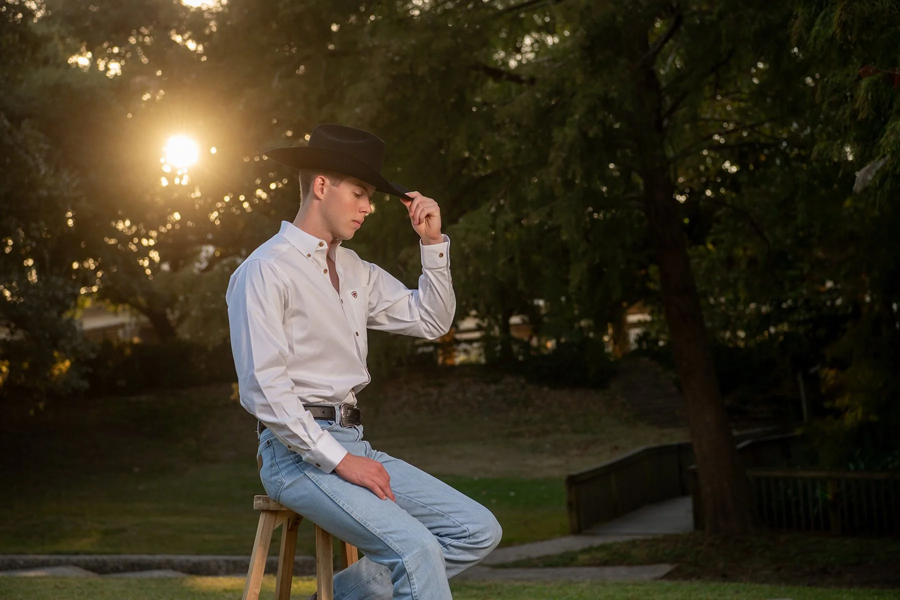 A young man sitting on a wooden stool outdoors during sunset, wearing a white shirt, blue jeans, and a black cowboy hat, with his hand adjusting the hat and a contemplative expression.