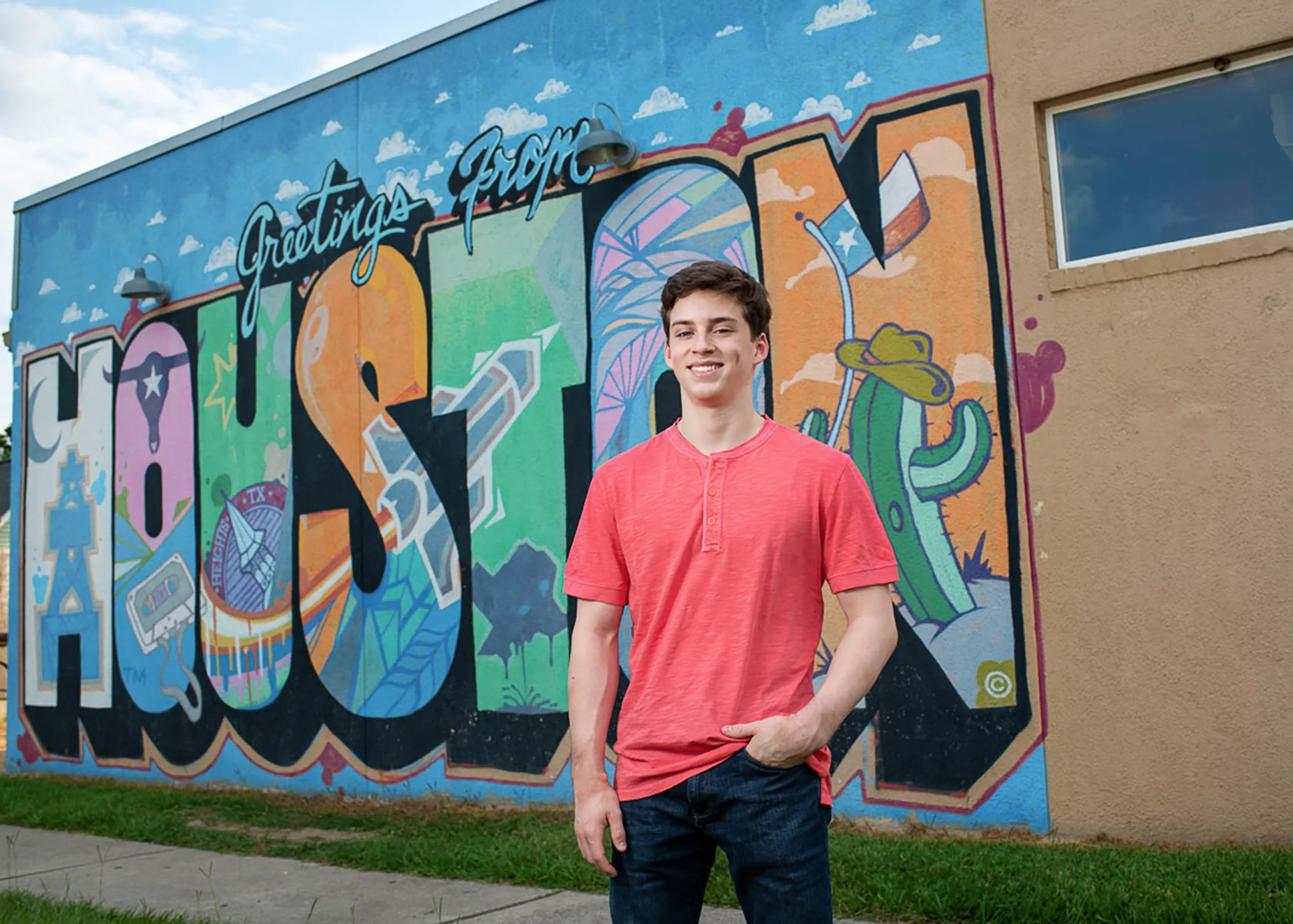 Young man in a red shirt standing in front of a colorful mural that reads "Greetings from Austin, TX" with various illustrations including clouds, a cactus, and abstract designs.