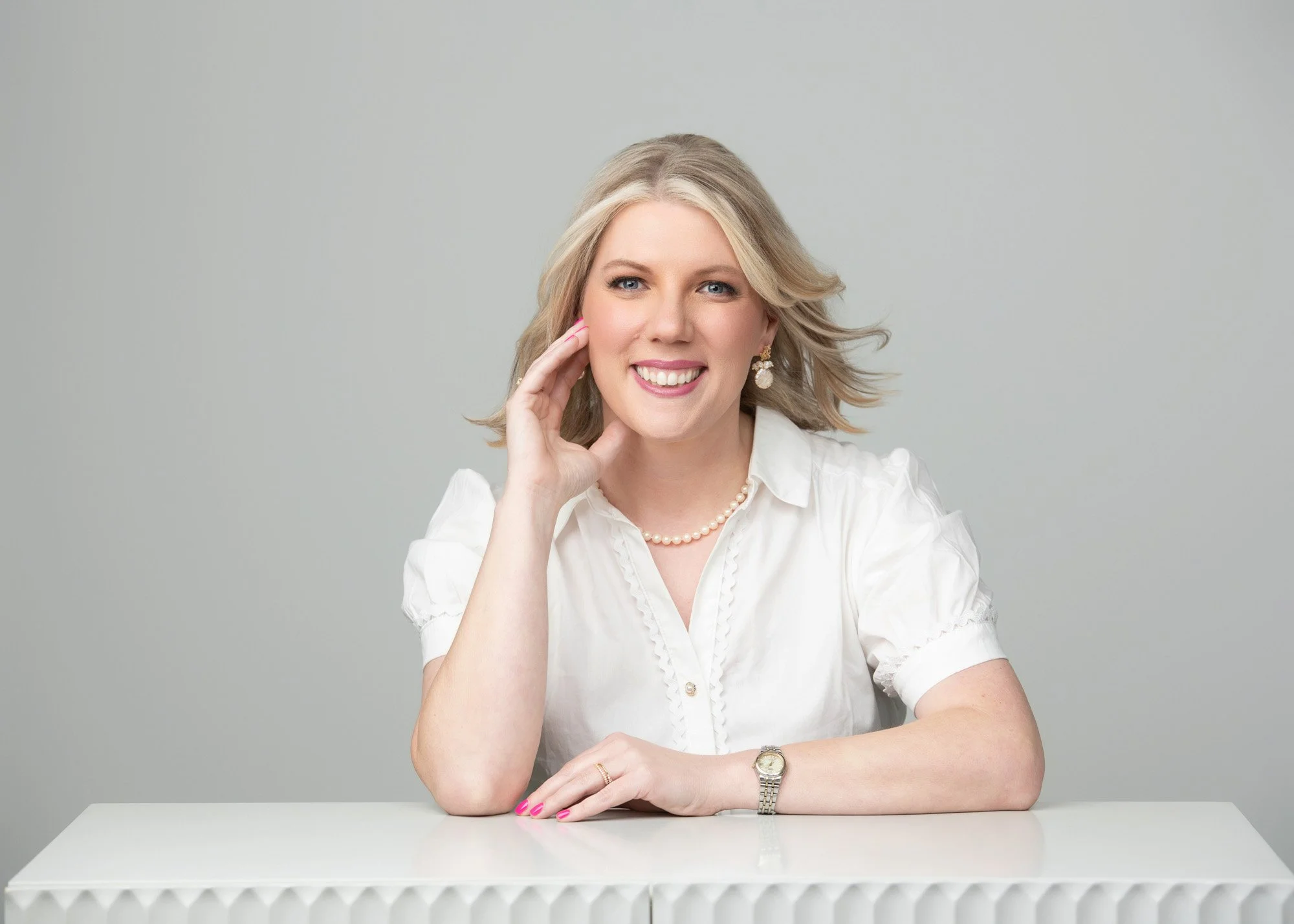 A woman with blonde hair smiling, sitting at a white table, touching her face with one hand, wearing a white blouse, pearl earrings, a pearl necklace, and a gold watch on her left wrist.