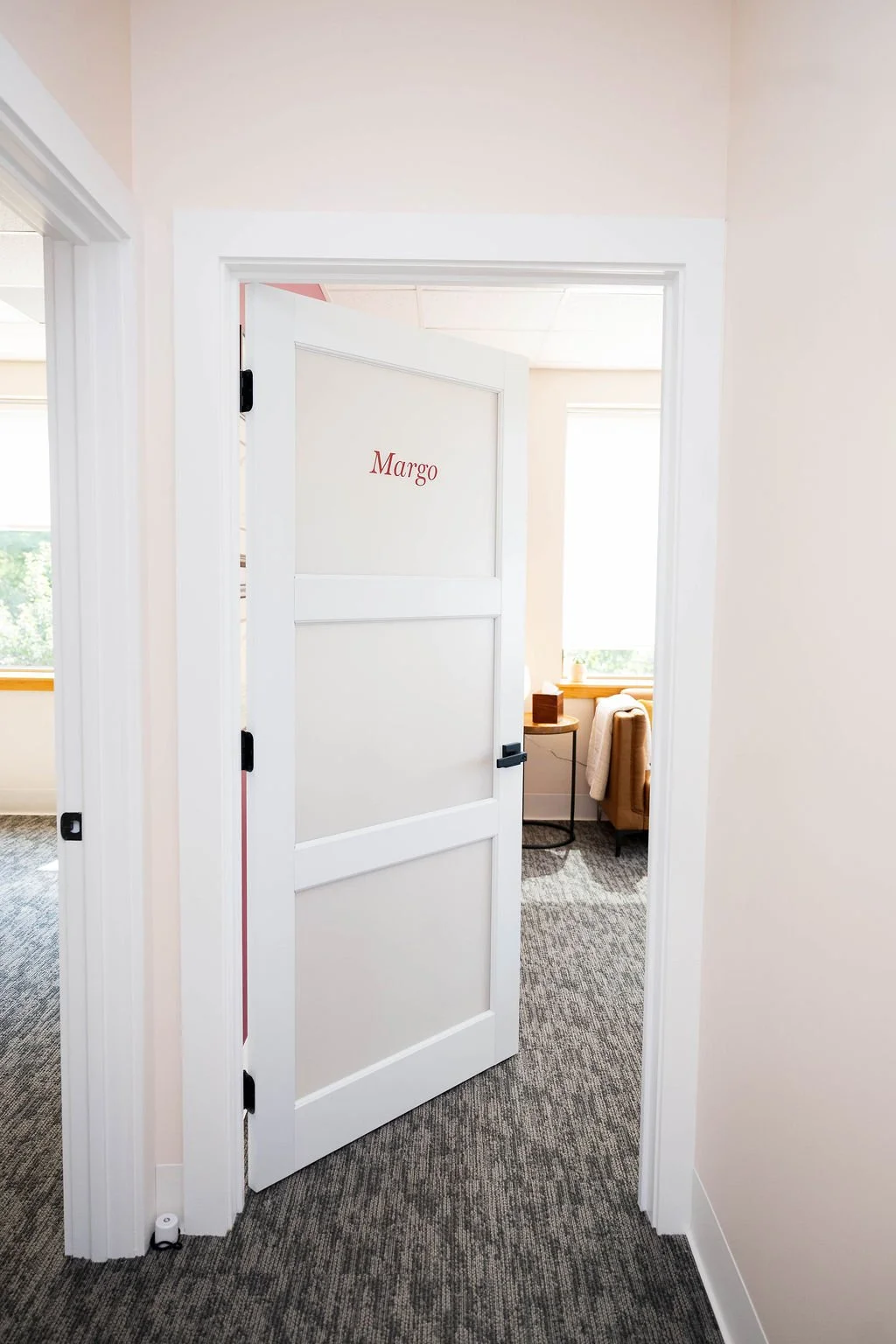 A white door slightly open with the name 'Margo' written in red on it, revealing a room with a window, a sofa, a side table, and a lamp.