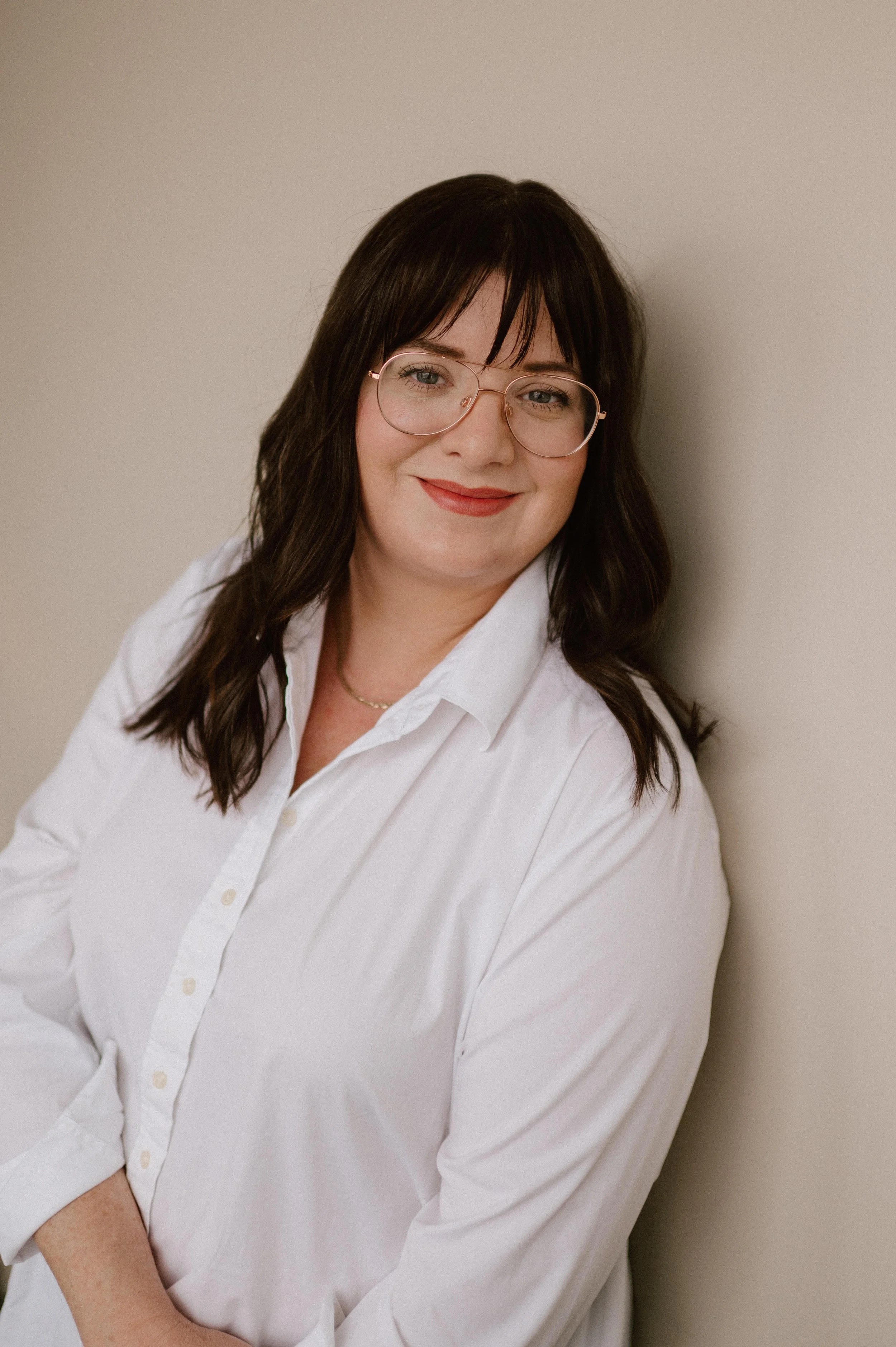 A smiling woman with dark brown hair and bangs leans against a neutral beige wall.  She is wearing clear-framed, gold-accented aviator glasses and a crisp white button-down shirt.  Her hair is styles in loose waves, looking directly at camera.