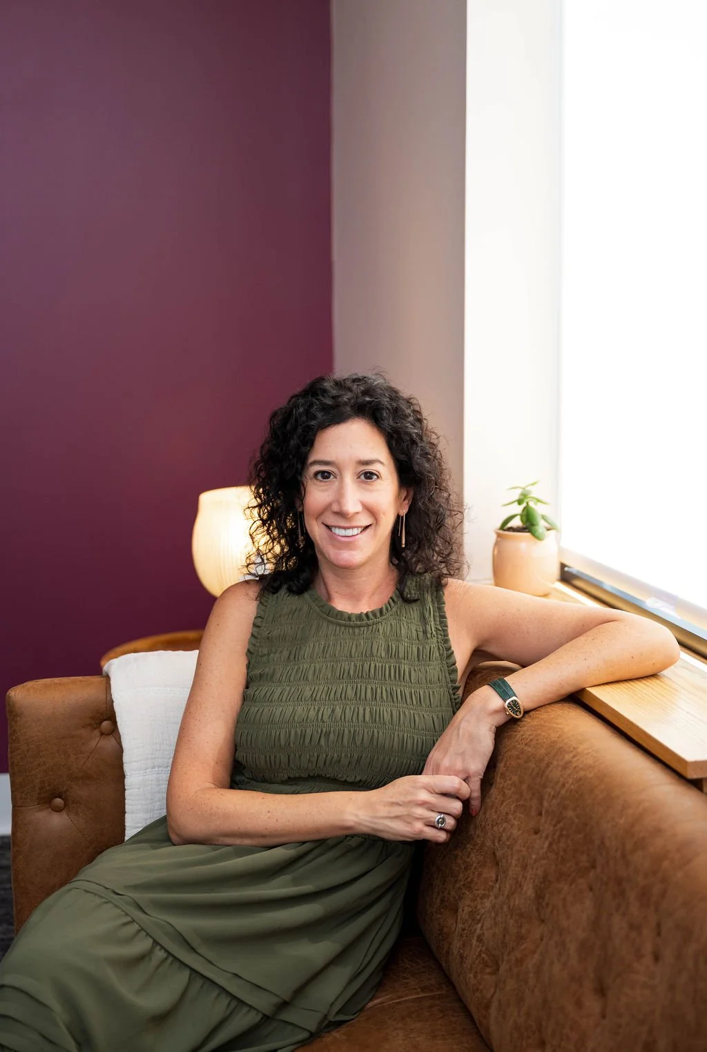 A woman with curly dark hair smiling, wearing a sleeveless green top, seated on a brown sofa in a room with a purple and beige wall background, a white cushion, a lamp, and a small green plant on the windowsill.
