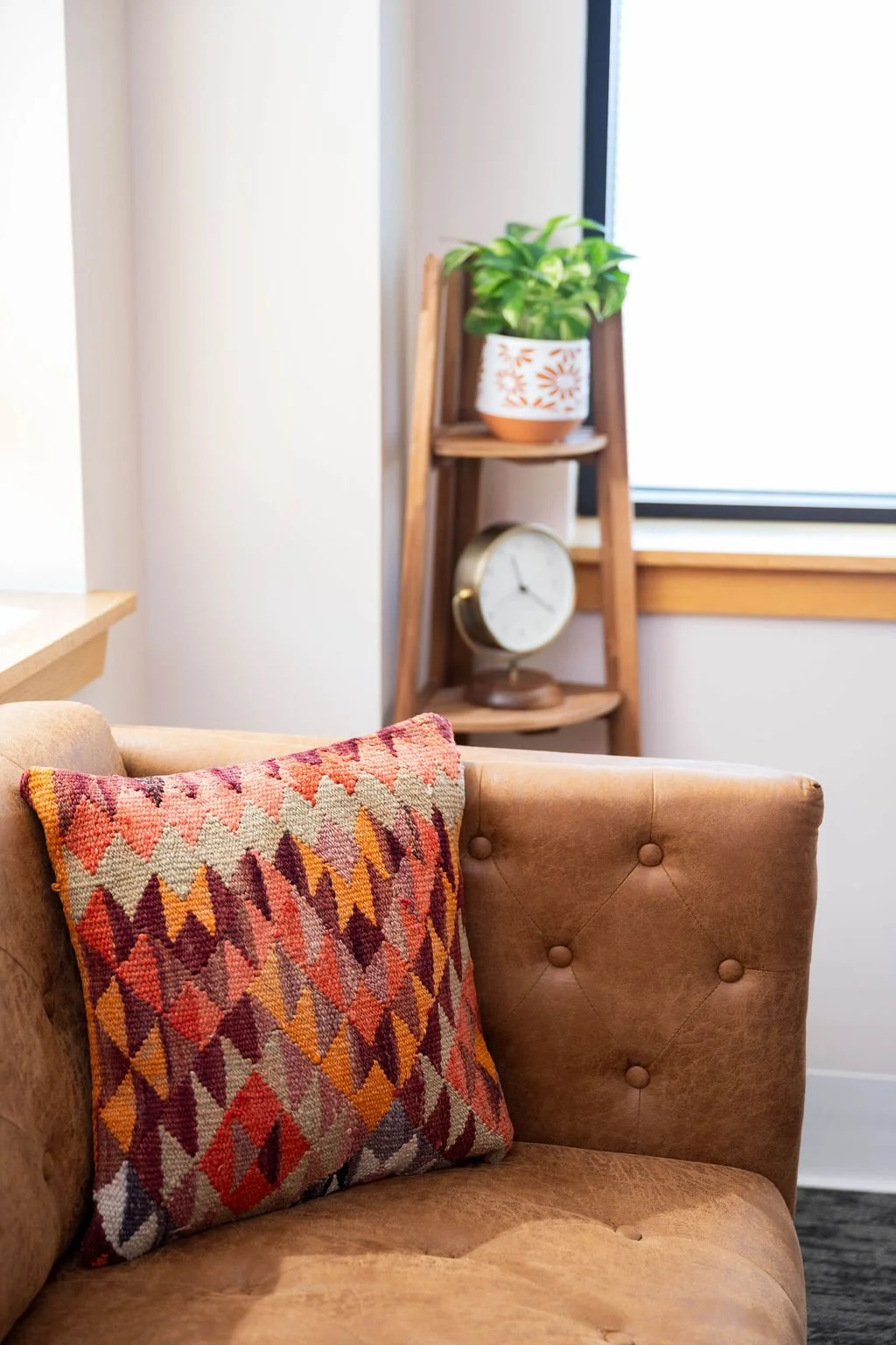 A living room with a beige tufted sofa, a colorful diamond-patterned throw pillow, a wooden ladder-style shelf with a potted plant, a vintage clock, and a window with wooden trim.