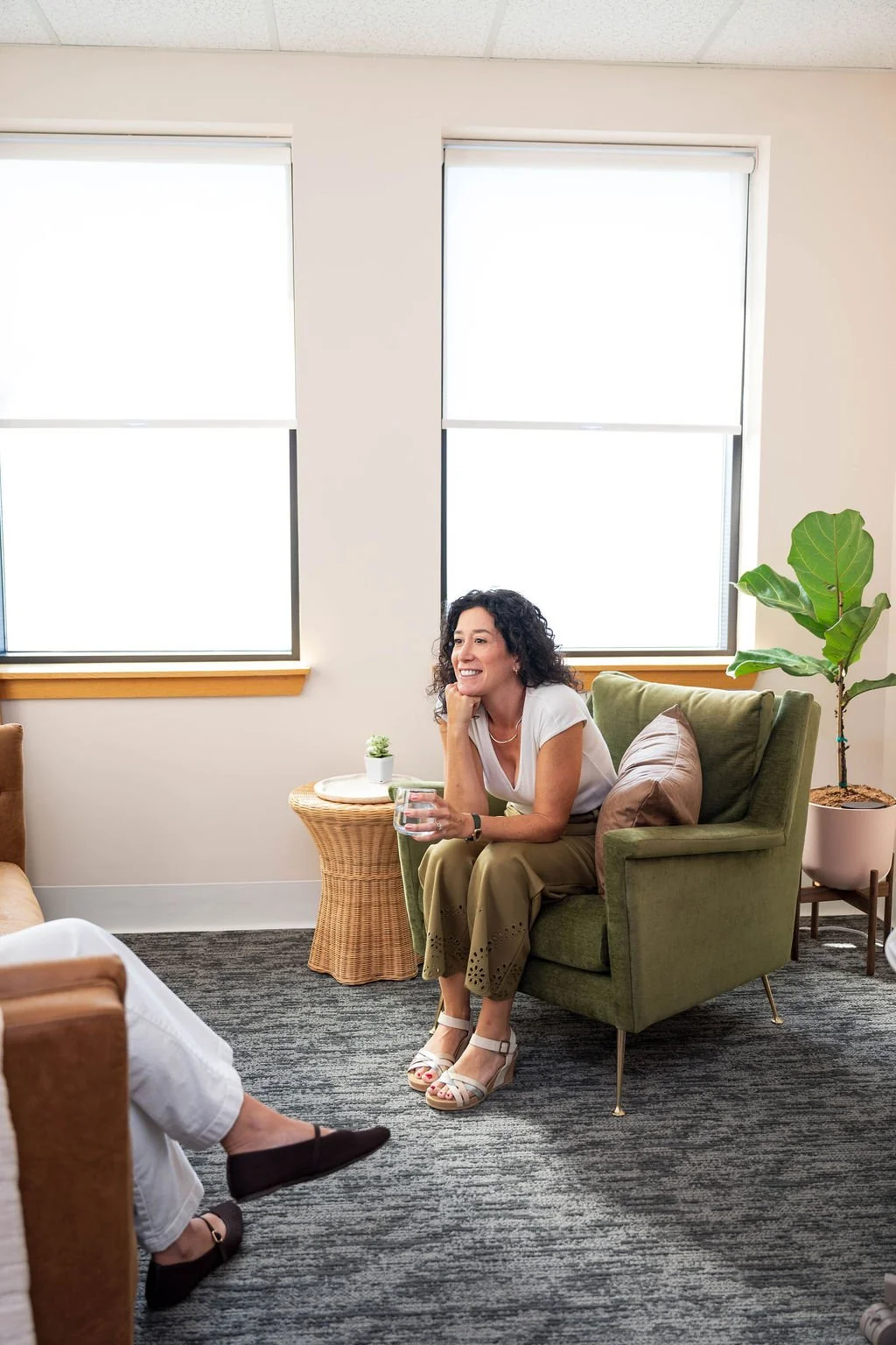A woman with curly dark hair sitting in a green armchair holding a glass of water, smiling and talking to another person whose legs are visible in the foreground. The room has light-colored walls, large windows with white blinds, a potted plant, and a small round wicker side table with a small plant on it.