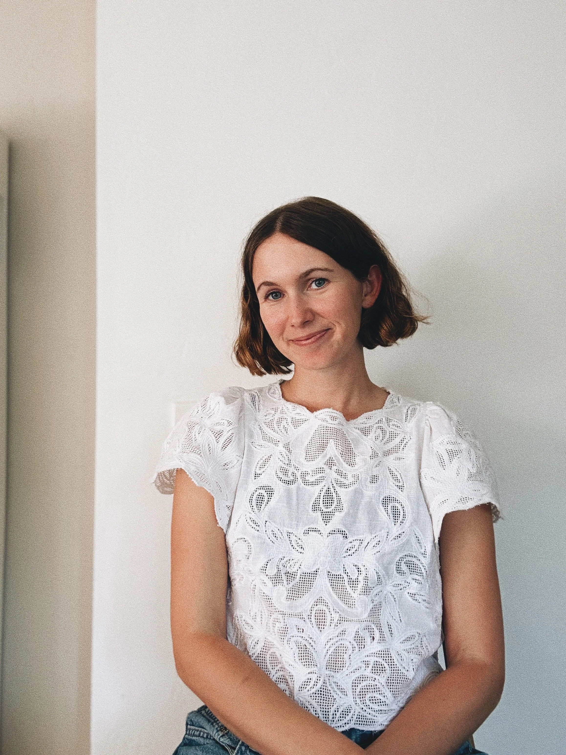 A  woman with a short brown bob haircut smiling gently at the camera. She is wearing an intricate white lace, short-sleeved blouse with cutout patterns, paired with blue denim jeans against a plain white wall background.