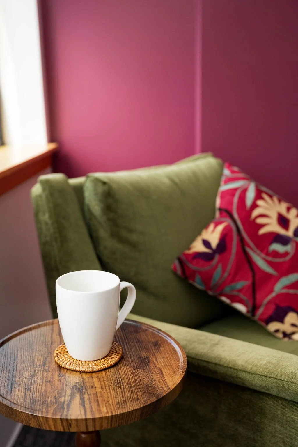 A cozy living room corner with a green velvet sofa, patterned throw pillows, a wooden side table, and a white ceramic mug on a woven coaster, set against a magenta wall.