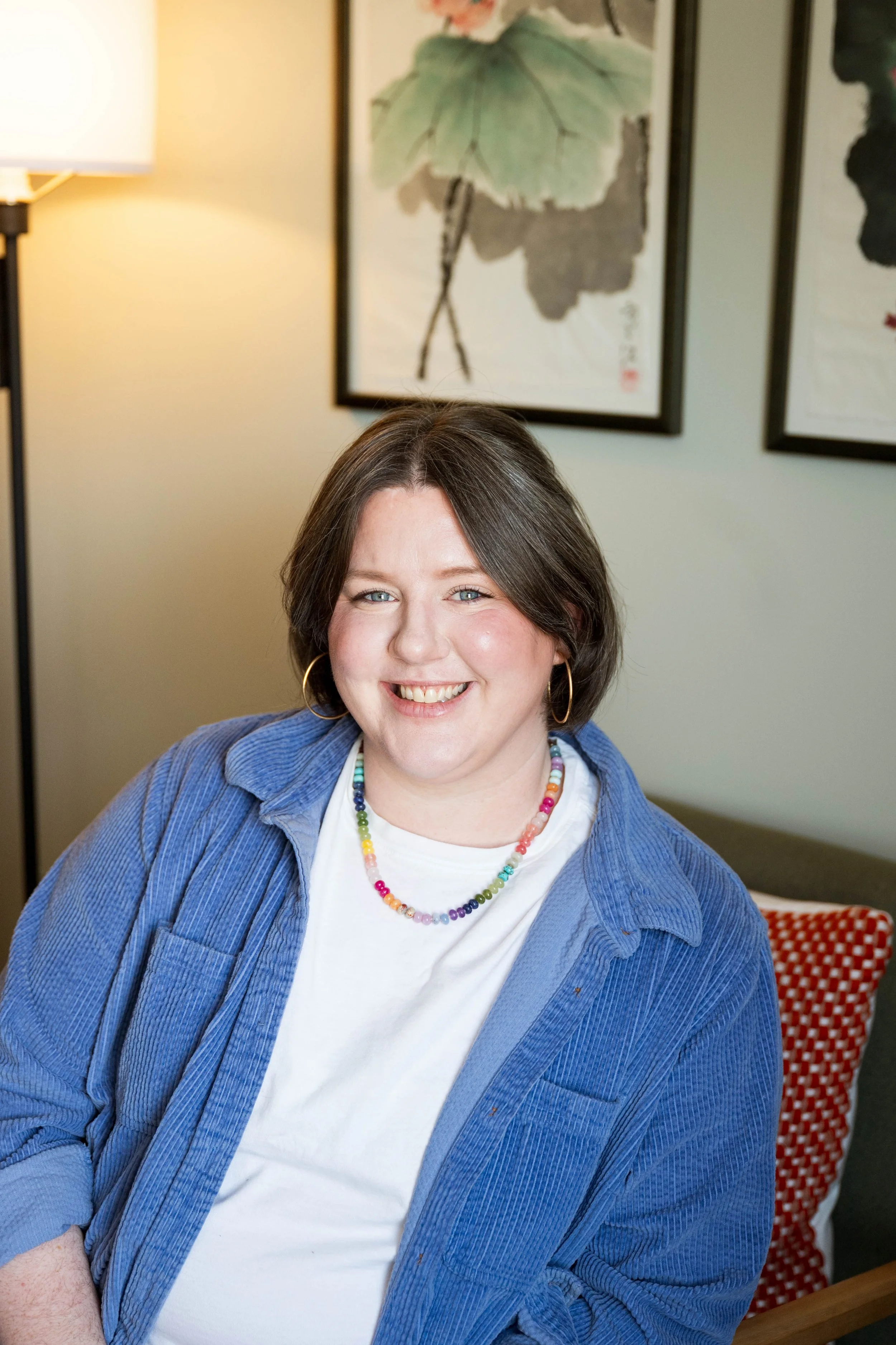 A smiling person with shoulder-length brown hair wearing a blue corduroy button-down shirt over a white t-shirt, large gold hoop earrings, and a colorful beaded necklace.