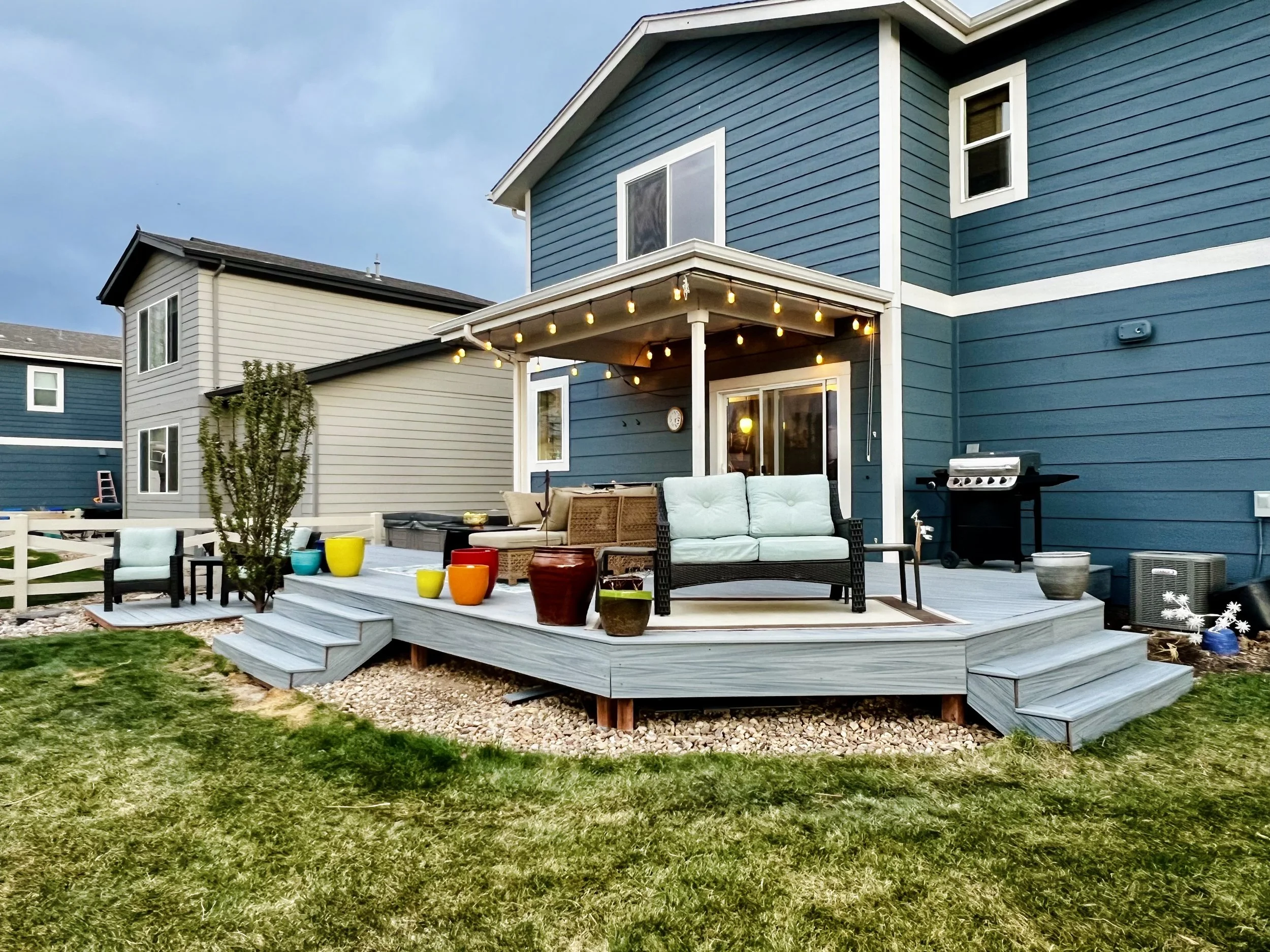 Backyard patio with outdoor furniture, colorful pots, and string lights in front of a blue house with white trim and sliding glass door.