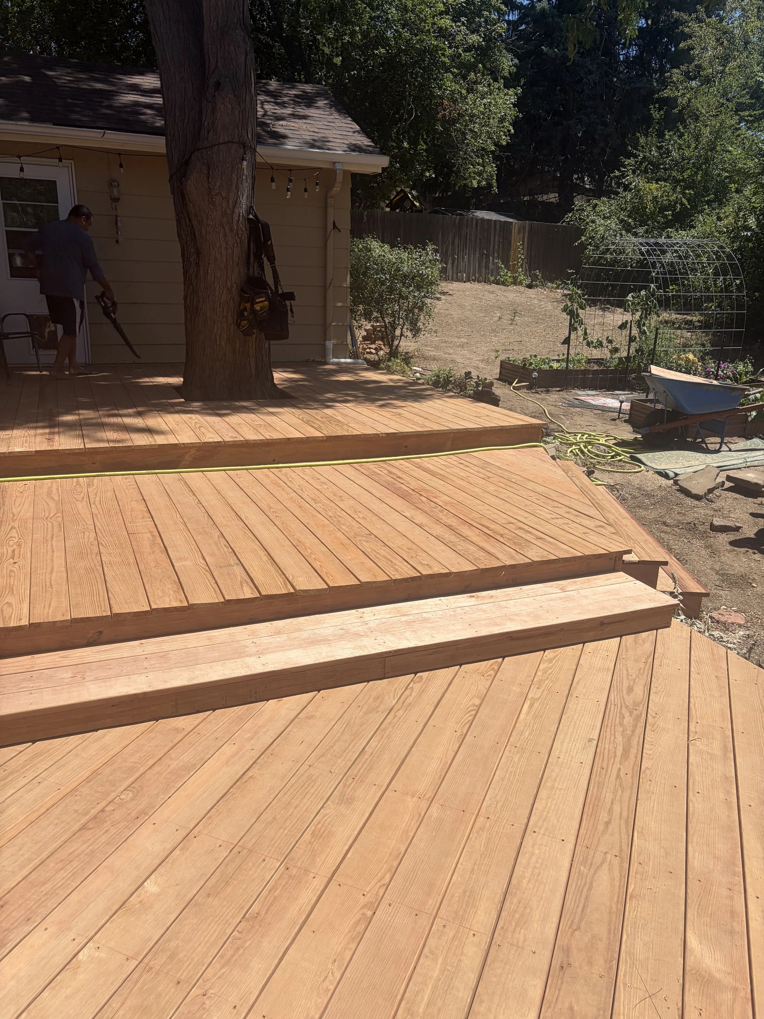 Backyard scene with a new wooden deck, a large tree, a person using a leaf blower, gardening tools, wheelbarrow, and a garden bed with plants.