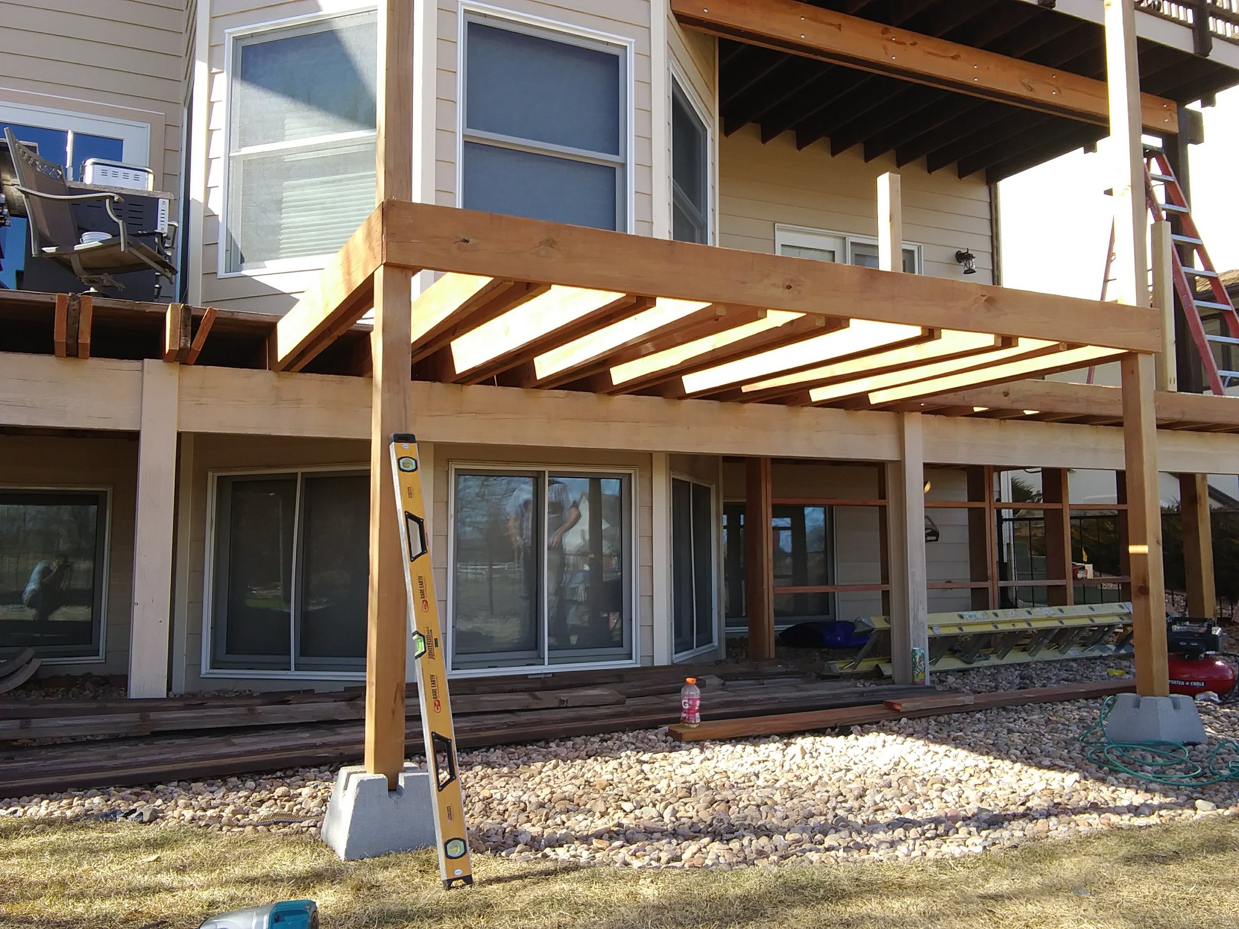 Back patio of a house under construction with scaffolding, a ladder, and construction tools visible. The house has a partially built deck with wooden beams and railings, large glass sliding doors, and exterior siding in beige color.