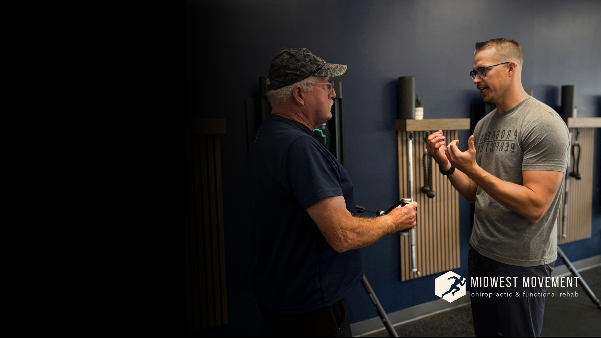 Two men engaged in conversation in a chiropractic clinic, standing in front of a wall with therapy tools, one wearing glasses and a gray t-shirt, the other with a camouflage hat and navy shirt.
